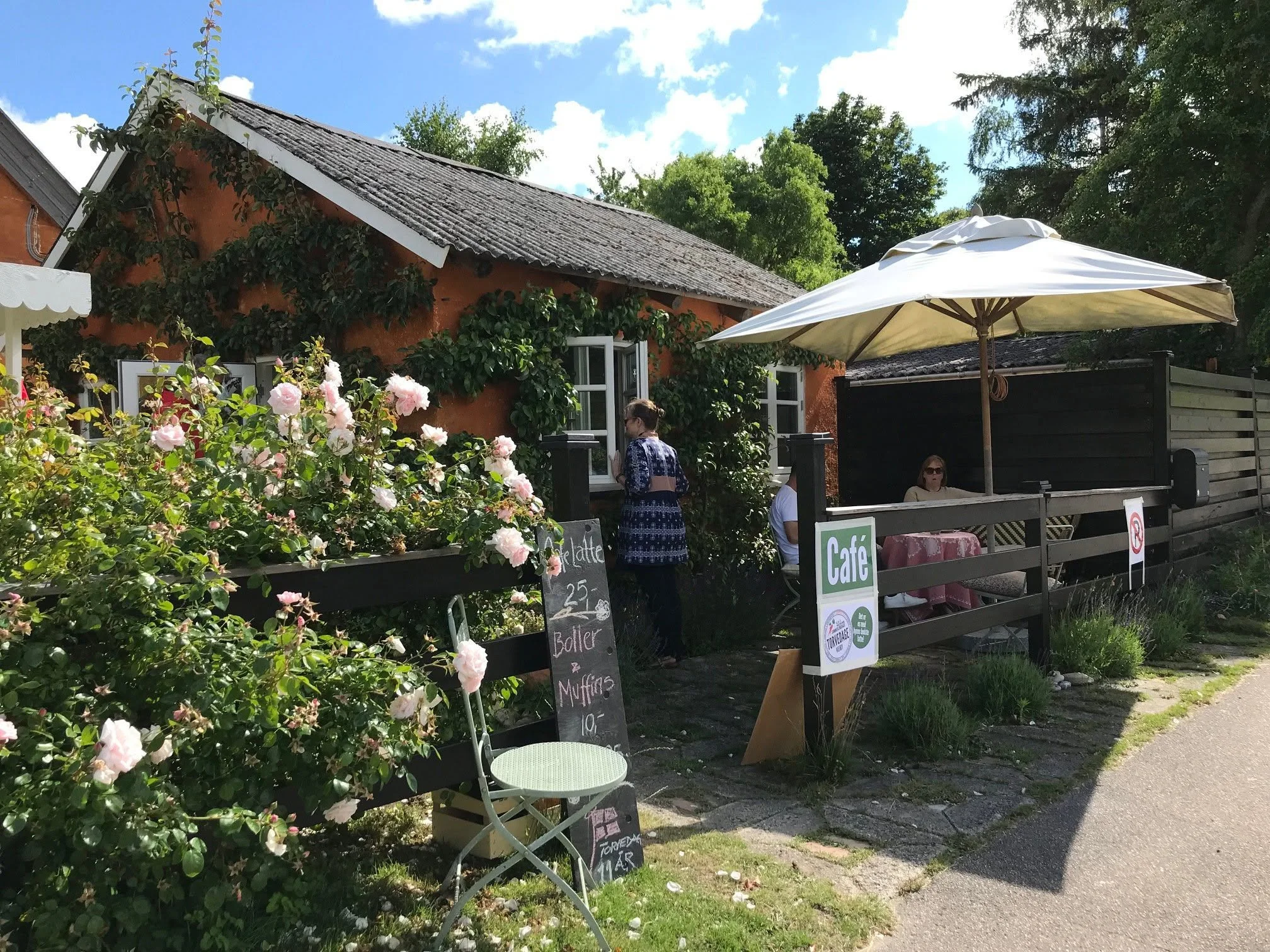 Et cafe-område med blomstrende planter og udendørs siddepladser under en stor parasol. Der er folk, der sidder og en person står ved en sort tavle med menukort.