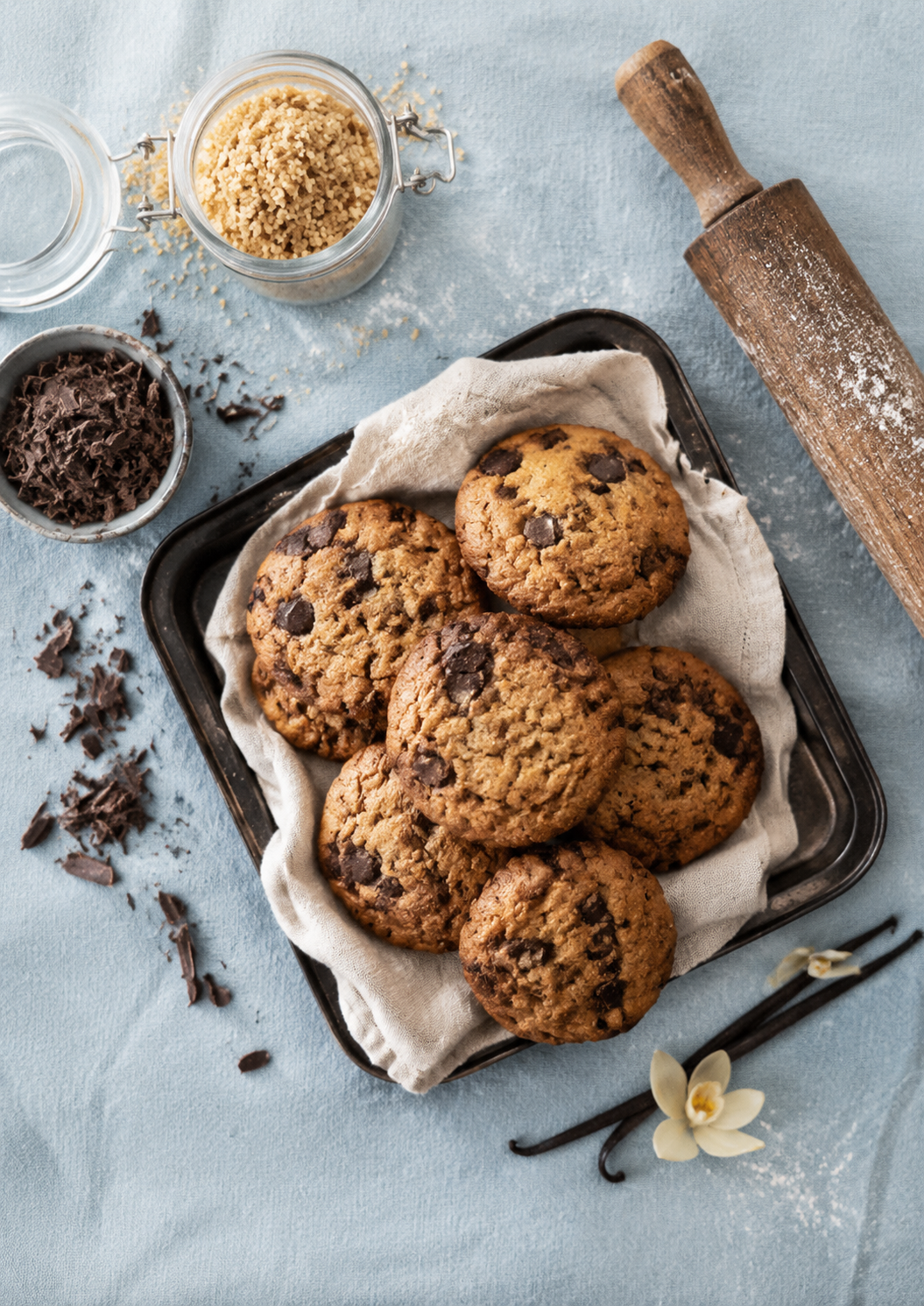 Friske, hjemmebagte chokoladechip cookies i en sort bakke med et stykke hvid klud. Der er en rulle til at bage kager og nogle vaniljeblade til pynt, samt chokoladestykker og sukker på et lyseblåt stof.