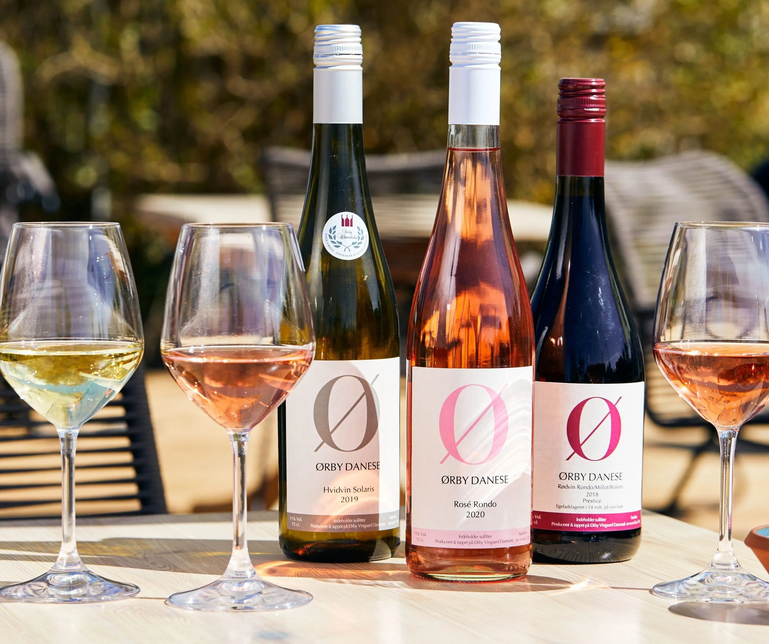 Tre flasker vin på et bord med tre tomme vin glas foran. Flaskerne har etiketter fra Ørby Danese vinproducenter.