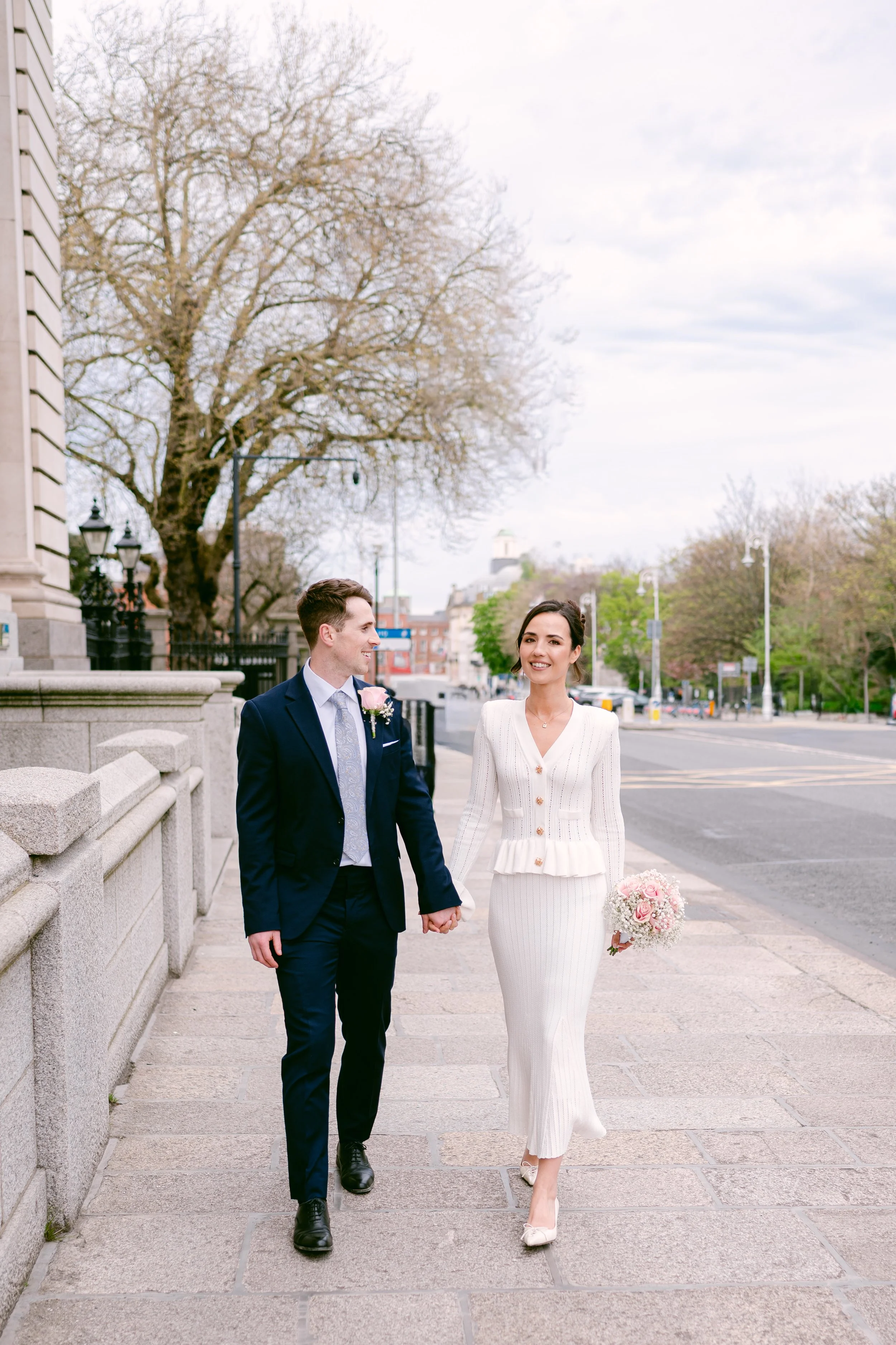 A bride and groom are walking hand in hand on a sidewalk outdoors in a city setting. The bride is wearing a white, long-sleeved wedding dress and holding a bouquet of pink and white flowers. The groom is dressed in a navy blue suit with a light shirt and tie. They are smiling and looking at each other.