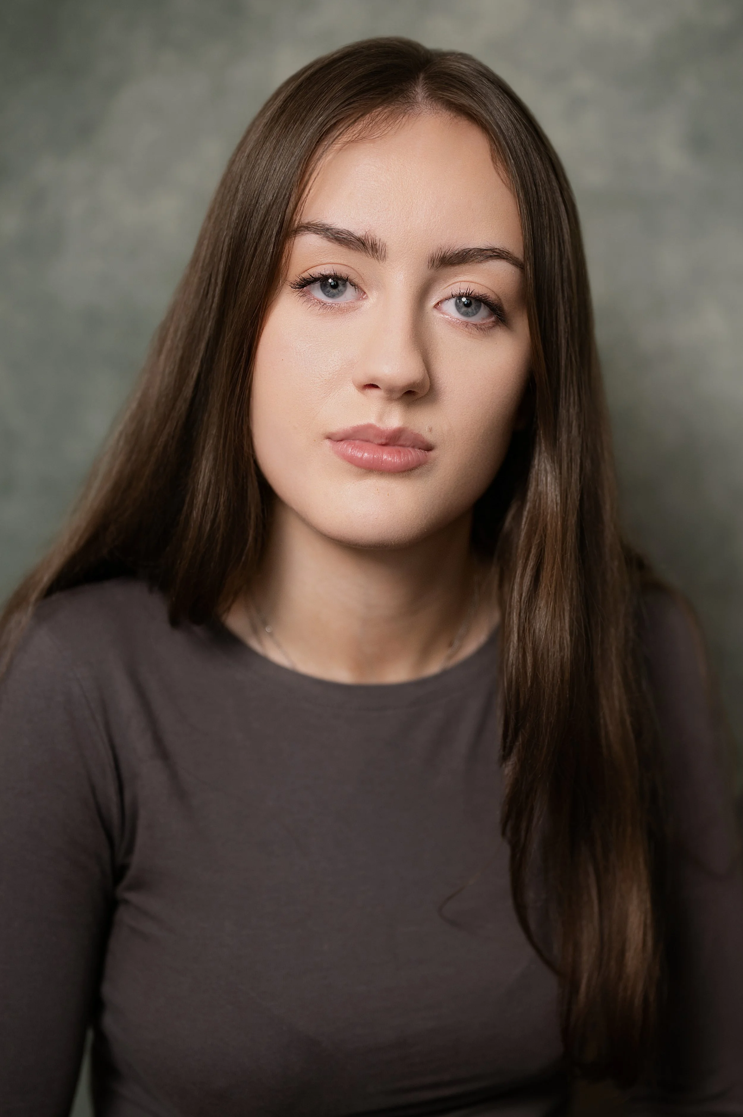 Close-up Actor Headshot of a young woman with long brown hair, light skin, and blue eyes, wearing a dark gray top, against a gray background.