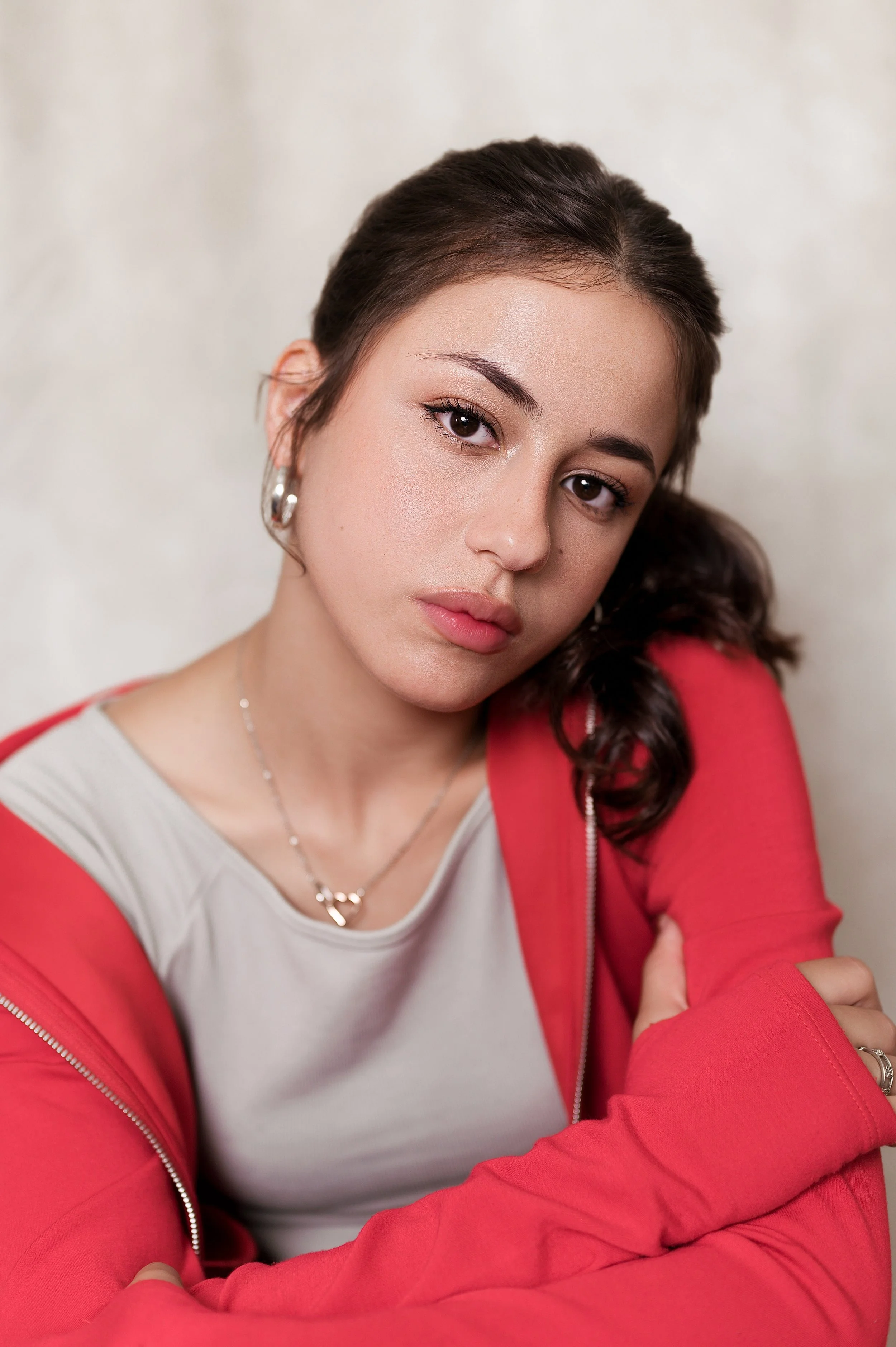 Close-up of a young woman with short brown hair, wearing silver hoop earrings, a light gray shirt, a silver necklace, and a red jacket. She has a neutral expression and is looking directly at the camera.