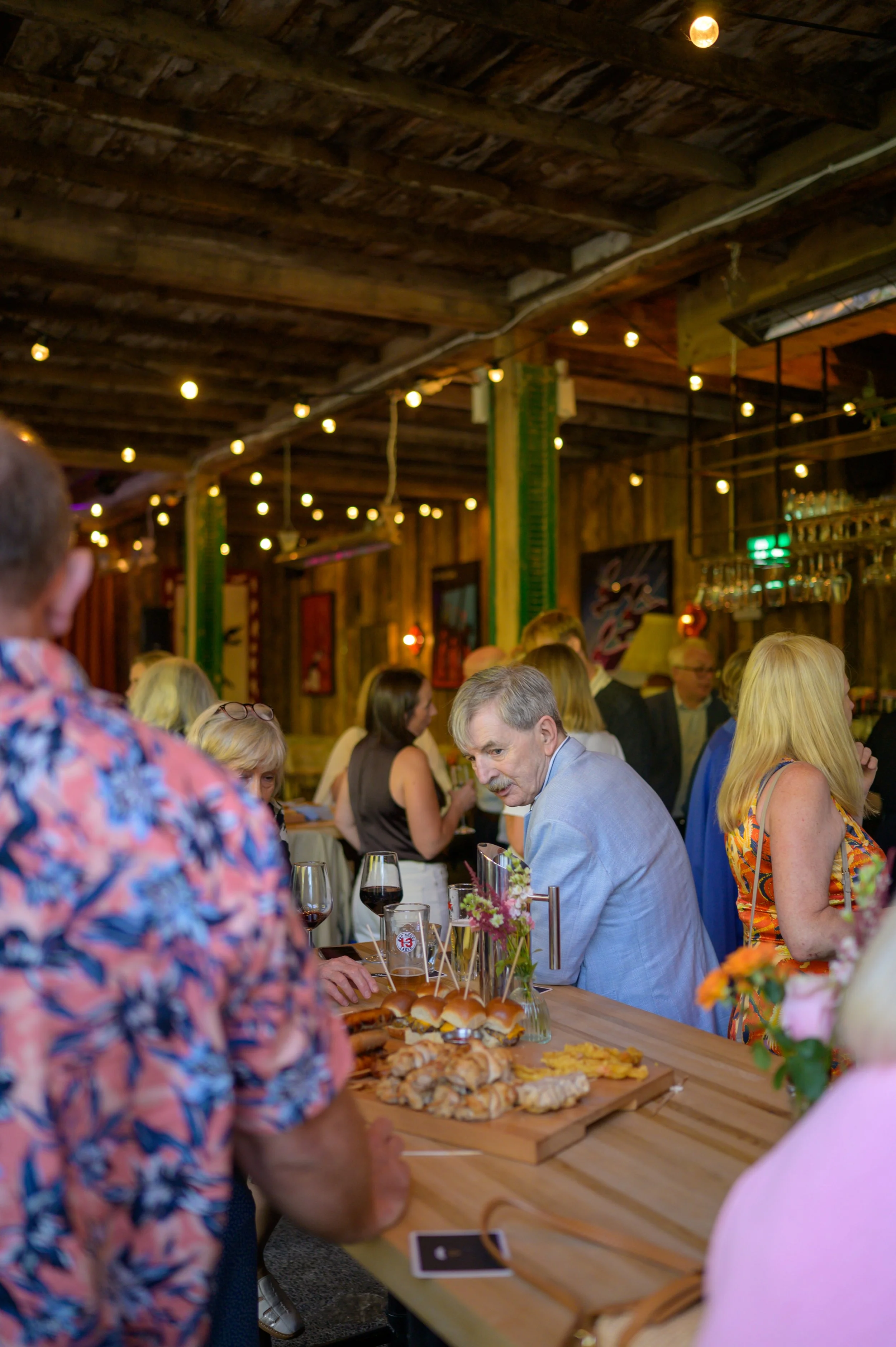 People gathered around a table with food and drinks in a rustic, wood-paneled room with string lights.