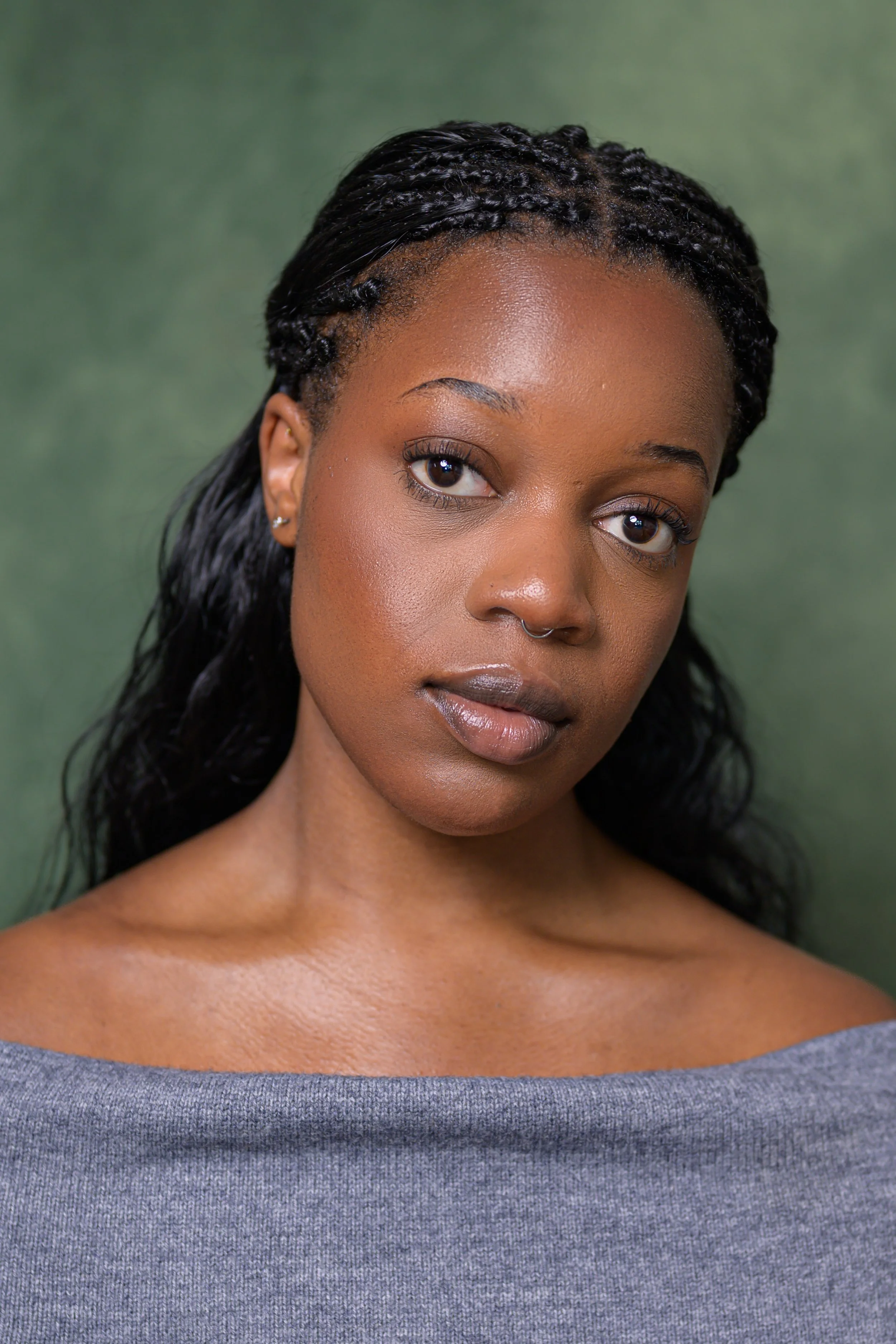 Close-up portrait of a young woman with dark braided hair, wearing a gray off-the-shoulder top, and looking slightly to the side against a green background.