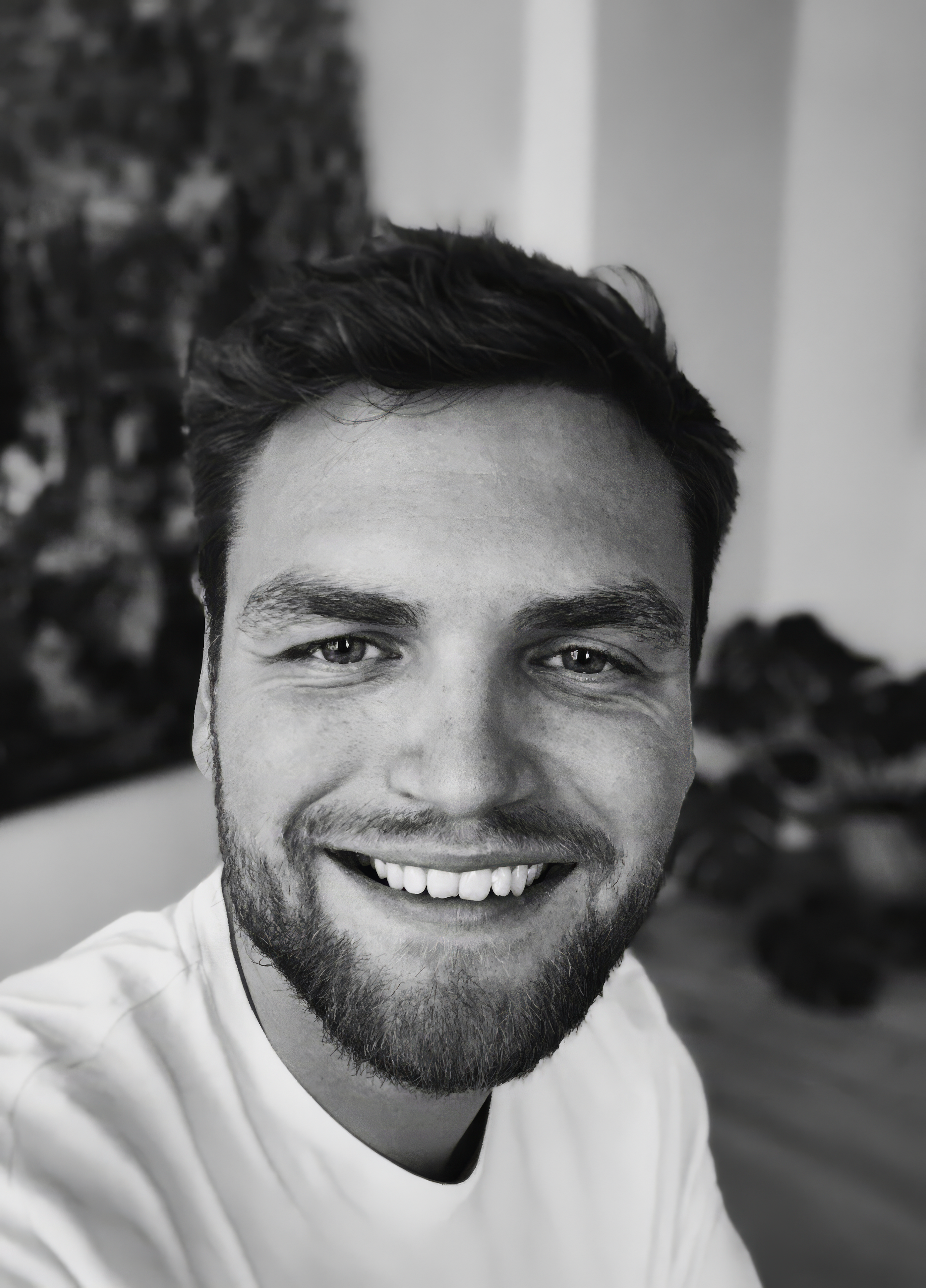 Et smilende ungt mand med kort mørkt hår og skæg, taget i selfie i sort-hvid, iført hvid t-shirt, baggrund med abstrakt vægdekoration.