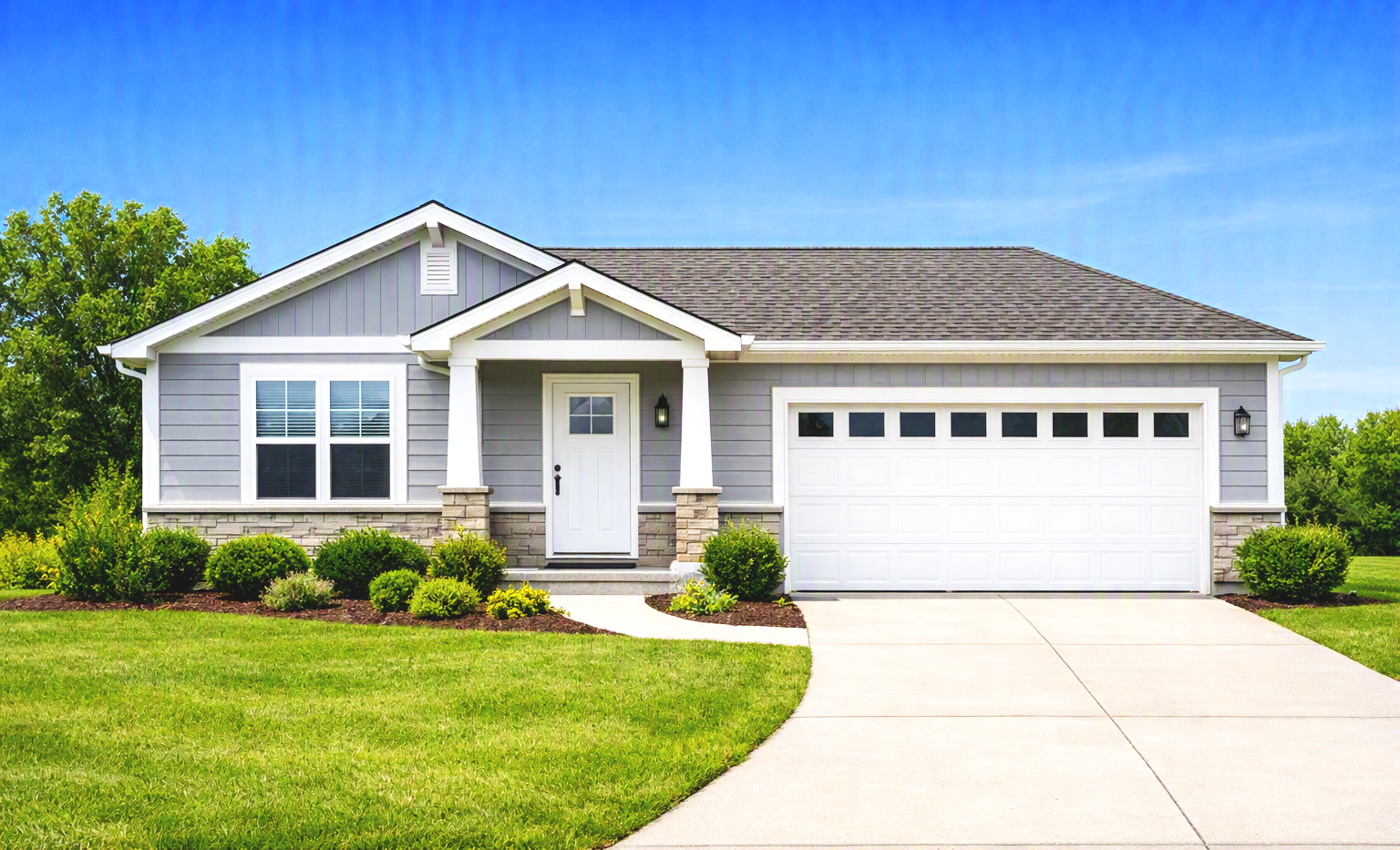 Front view of a modern suburban house with a gray exterior, white trim, two-car garage, small front porch with white door, landscaped yard, green lawn, and a bright sunny sky.
