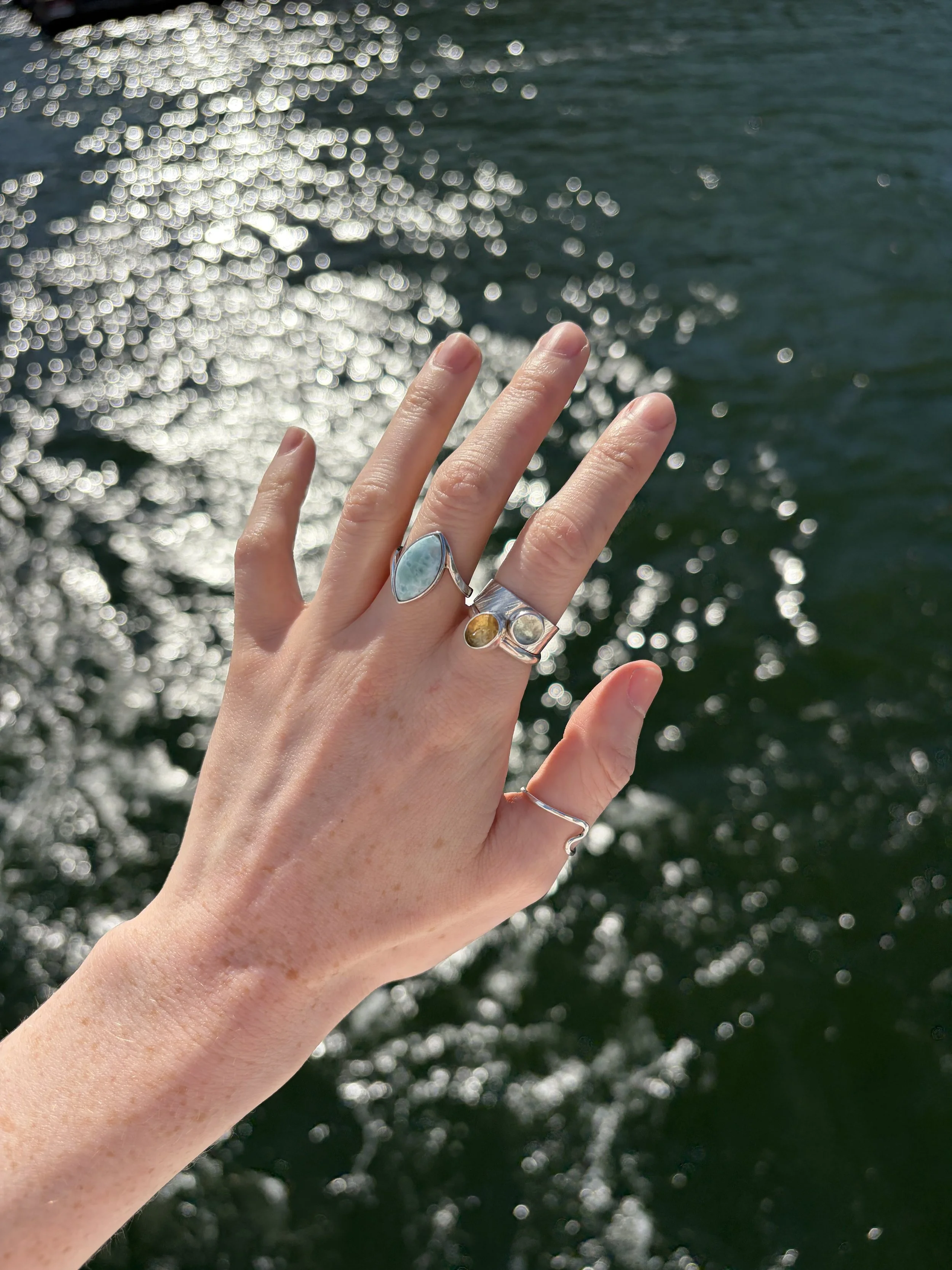Close-up of a person's hand with rings, held against a background of sparkling water with sunlight reflecting off it.