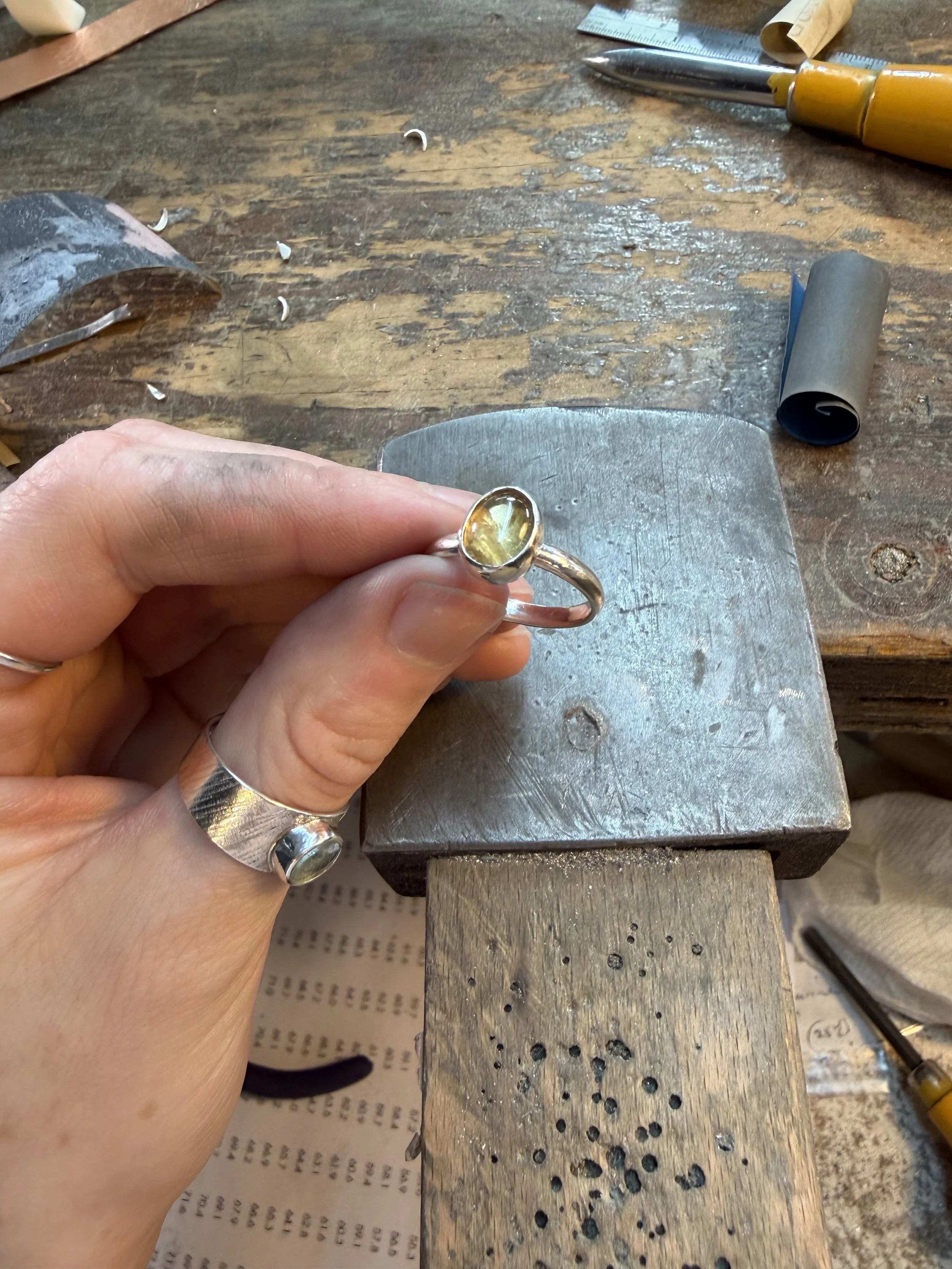 A jeweler's workbench with a hand holding a ring with a citrine gemstone, jewelry tools, and various small metal shavings and pieces on the wooden surface. Canada. Jewellery.  Silver Jewellery. Silver jewelry. 