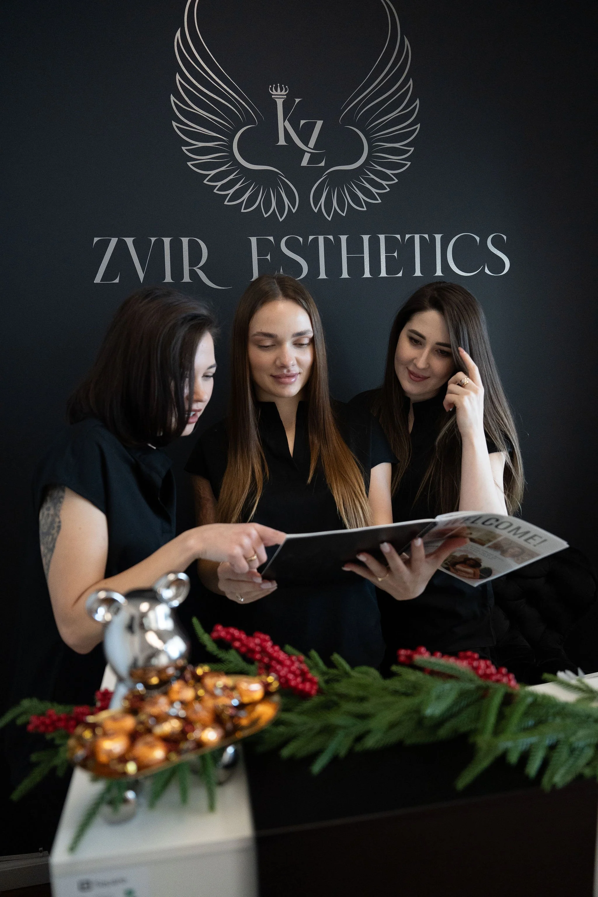 Three women dressed in black, looking at a magazine or brochure together inside a wellness or aesthetic clinic. The background features a black wall with the gold and white logo of 'Zvir Esthetics' with winged design.