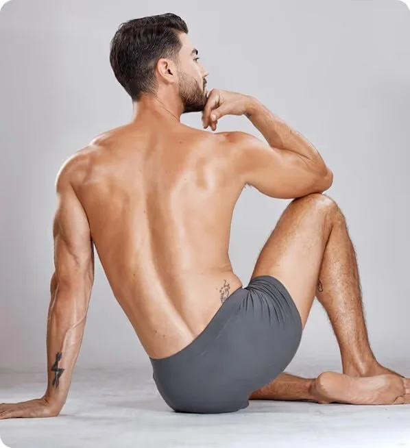A shirtless man with a beard and tattoos, sitting on the floor with one leg raised, in a contemplative pose against a plain background.