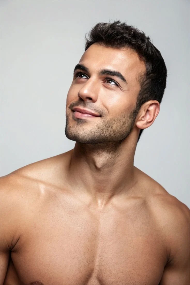 A shirtless young man with short dark hair and light skin, looking upward with a slight smile, against a plain light gray background.