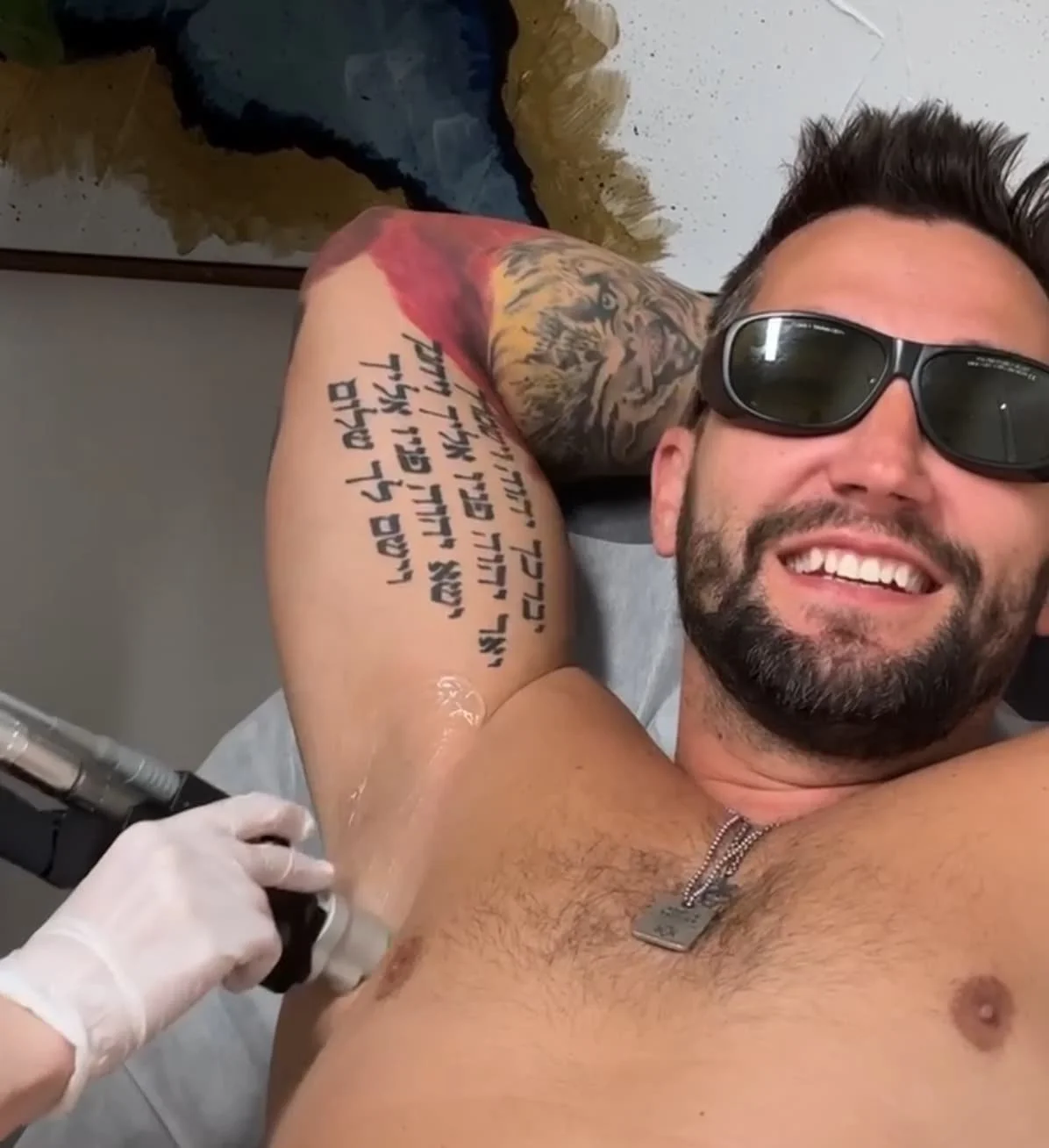 A smiling man with sunglasses lying shirtless on a medical chair, receiving a tattoo on his upper arm from a tattoo artist wearing gloves.