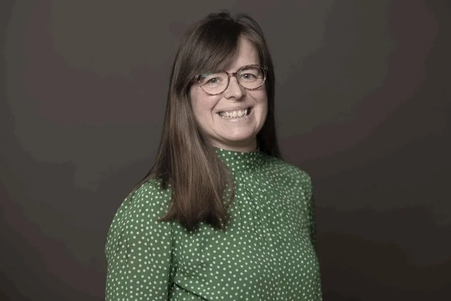 A woman with long brown hair wearing glasses and a green polka dot blouse, smiling against a dark background.