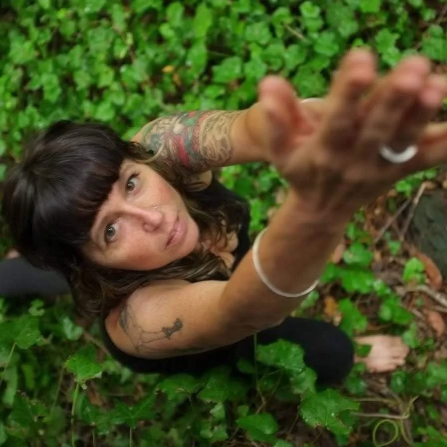 A yogini looks up at the camera while in Warrior 1 pose, surrounded by green outdoor plants.