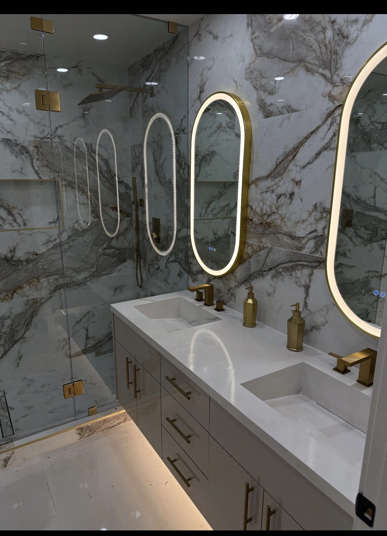 Modern bathroom with marble walls, dual sinks, gold faucets, illuminated mirrors, and a glass shower enclosure with gold accents.