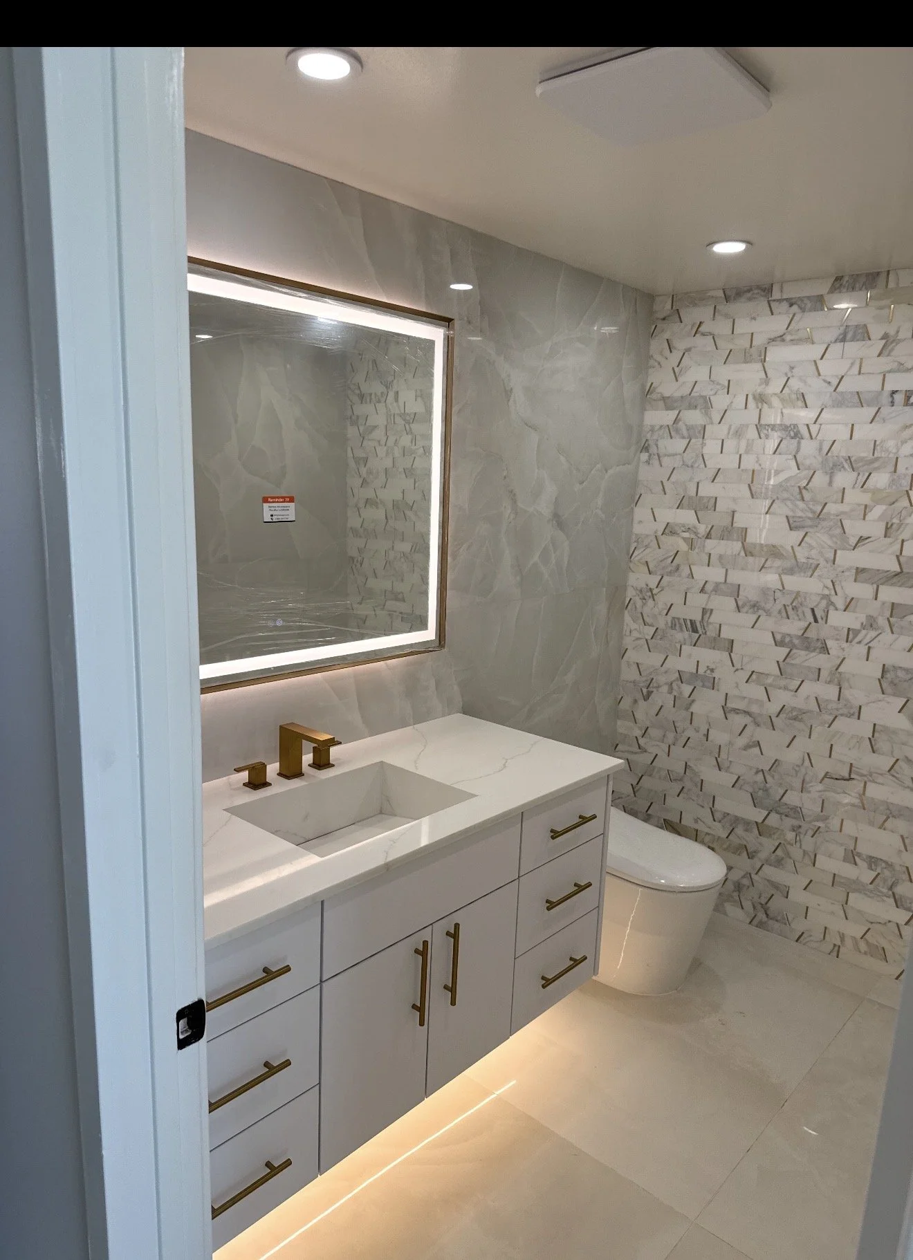 Modern bathroom with white vanity, gold hardware, rectangular mirror with backlighting, marble countertops, wood-look upload toilet, and textured tile wall.