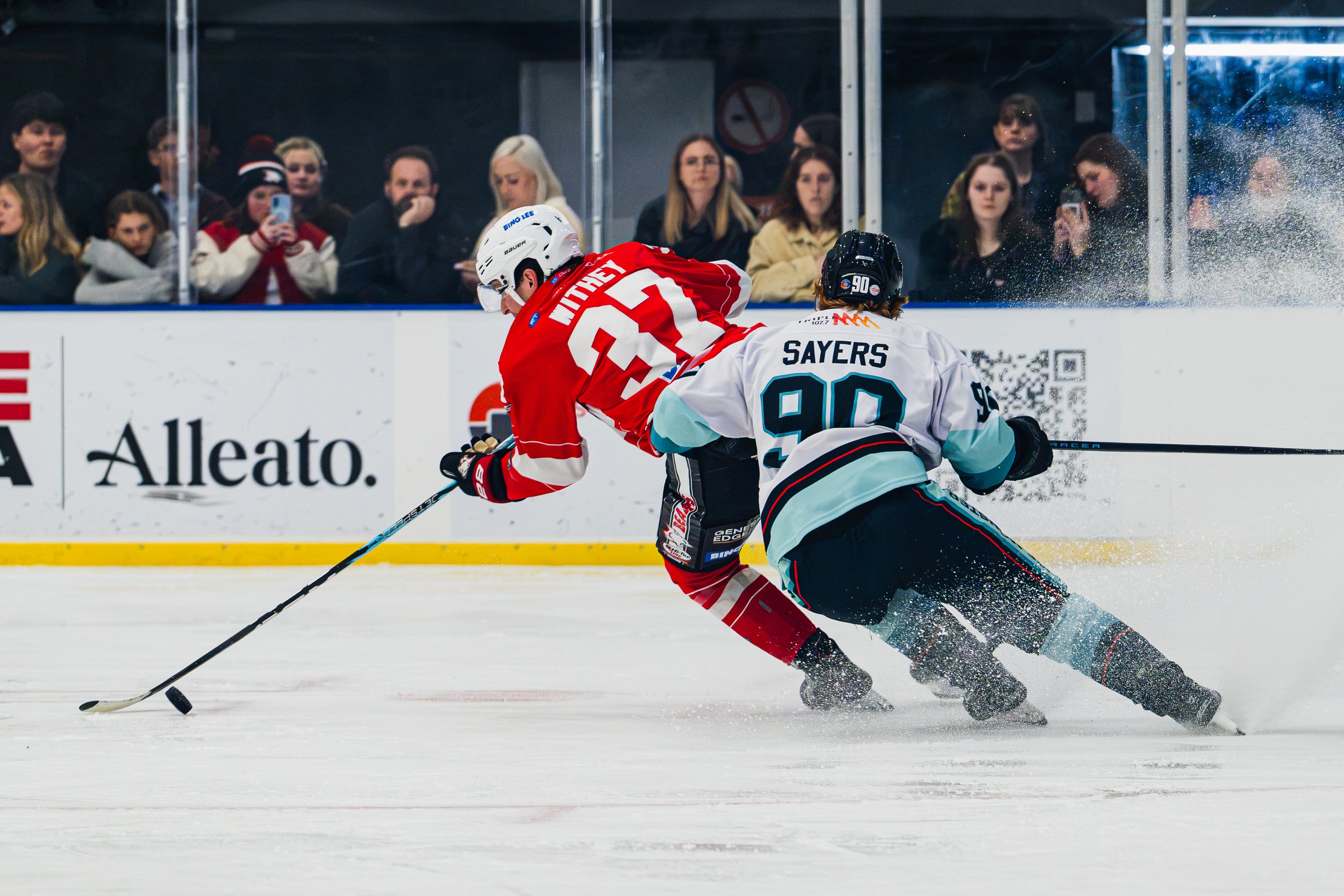 Two hockey players are racing for the puck on the ice rink, with one player in a red jersey and the other in a white jersey. Spectators watch from behind the glass, some taking photos or videos.