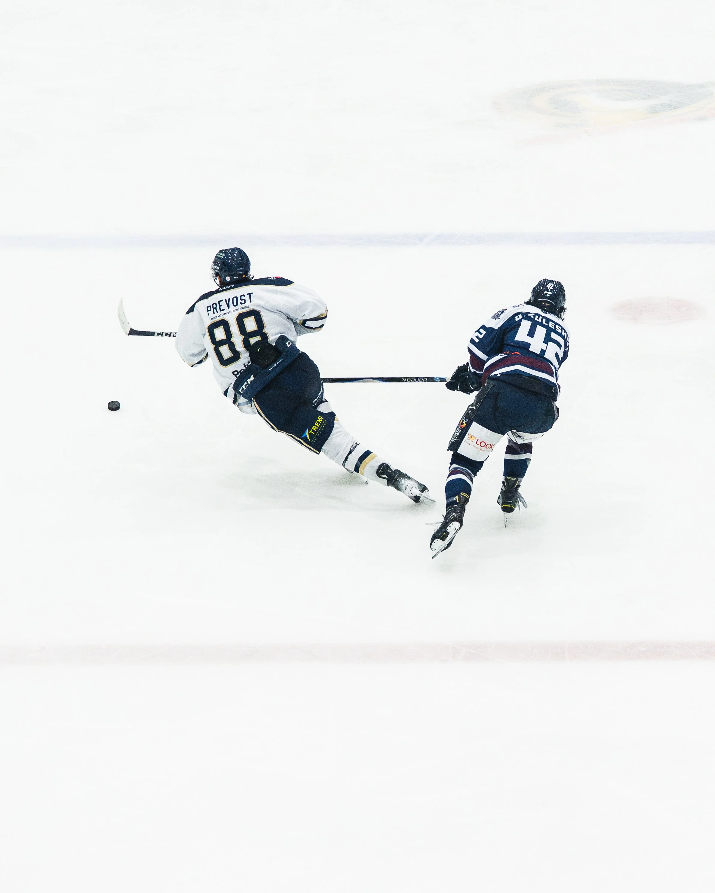 Two ice hockey players in action on the ice rink, one in a white jersey and the other in a dark jersey, skating towards the puck.