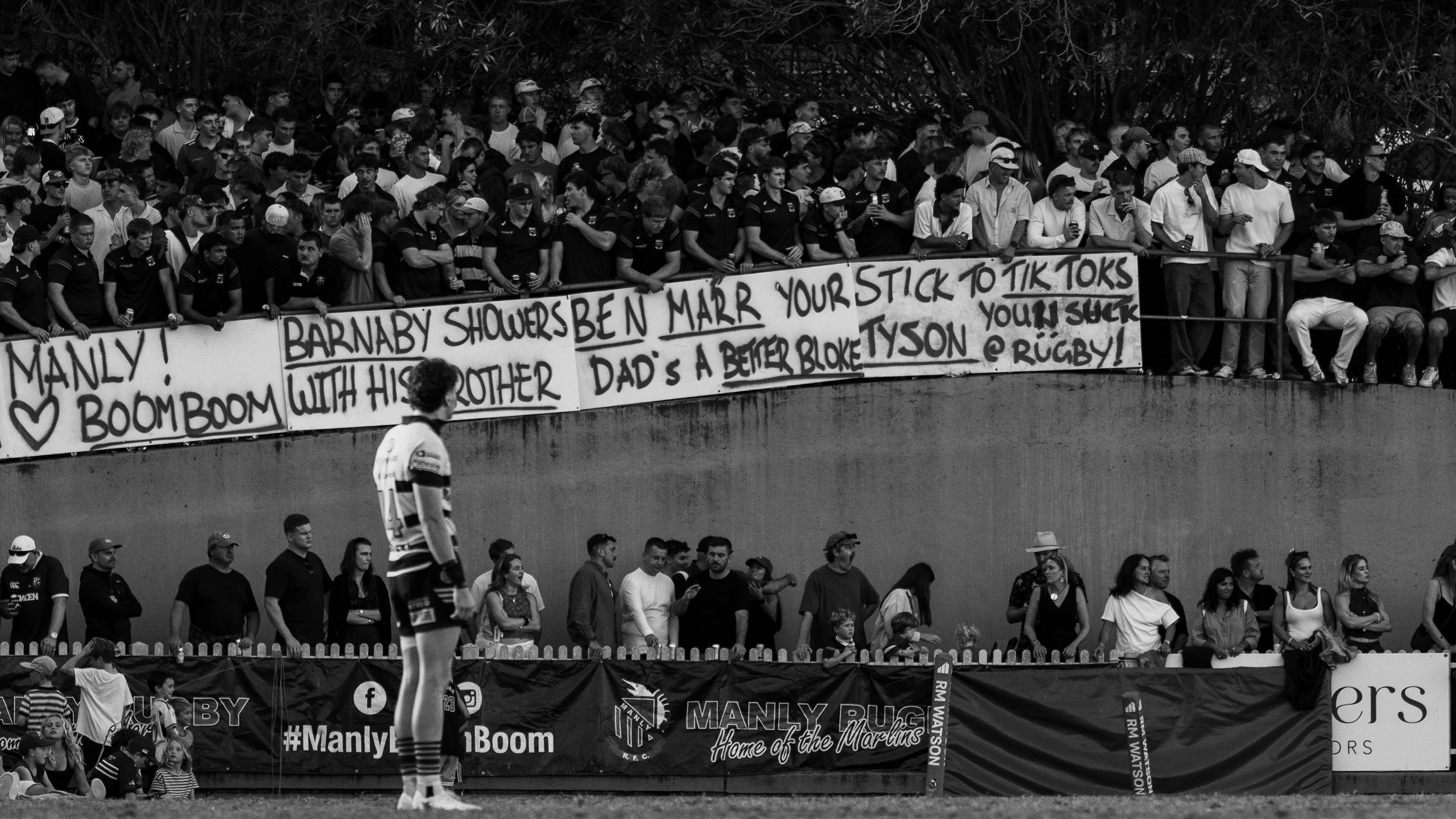 Black and white photo of a rugby match with a crowd of spectators sitting on a raised platform, holding signs and watching the game.