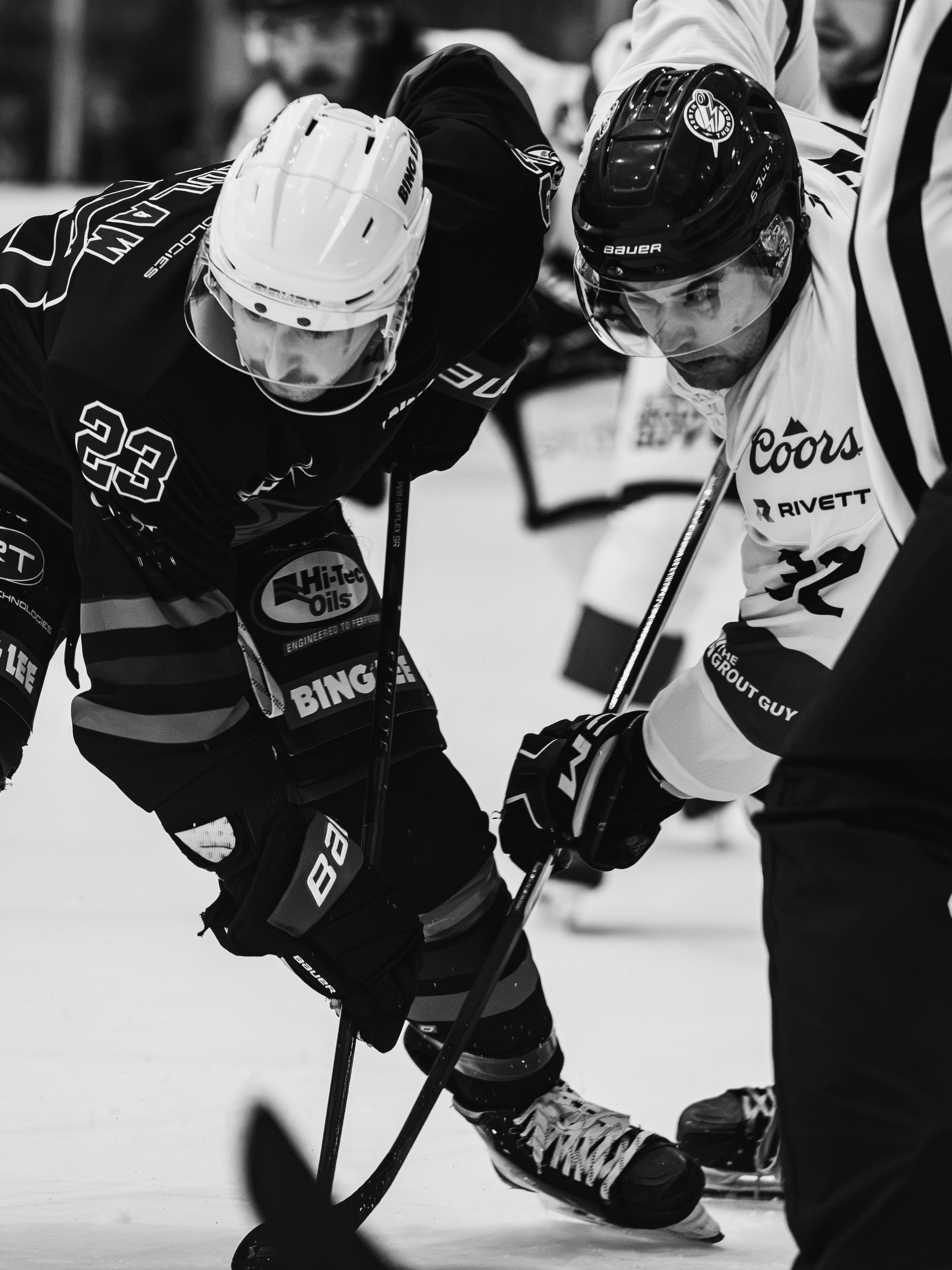 Two ice hockey players face off on the ice, wearing helmets and gear, with their sticks on the ice.