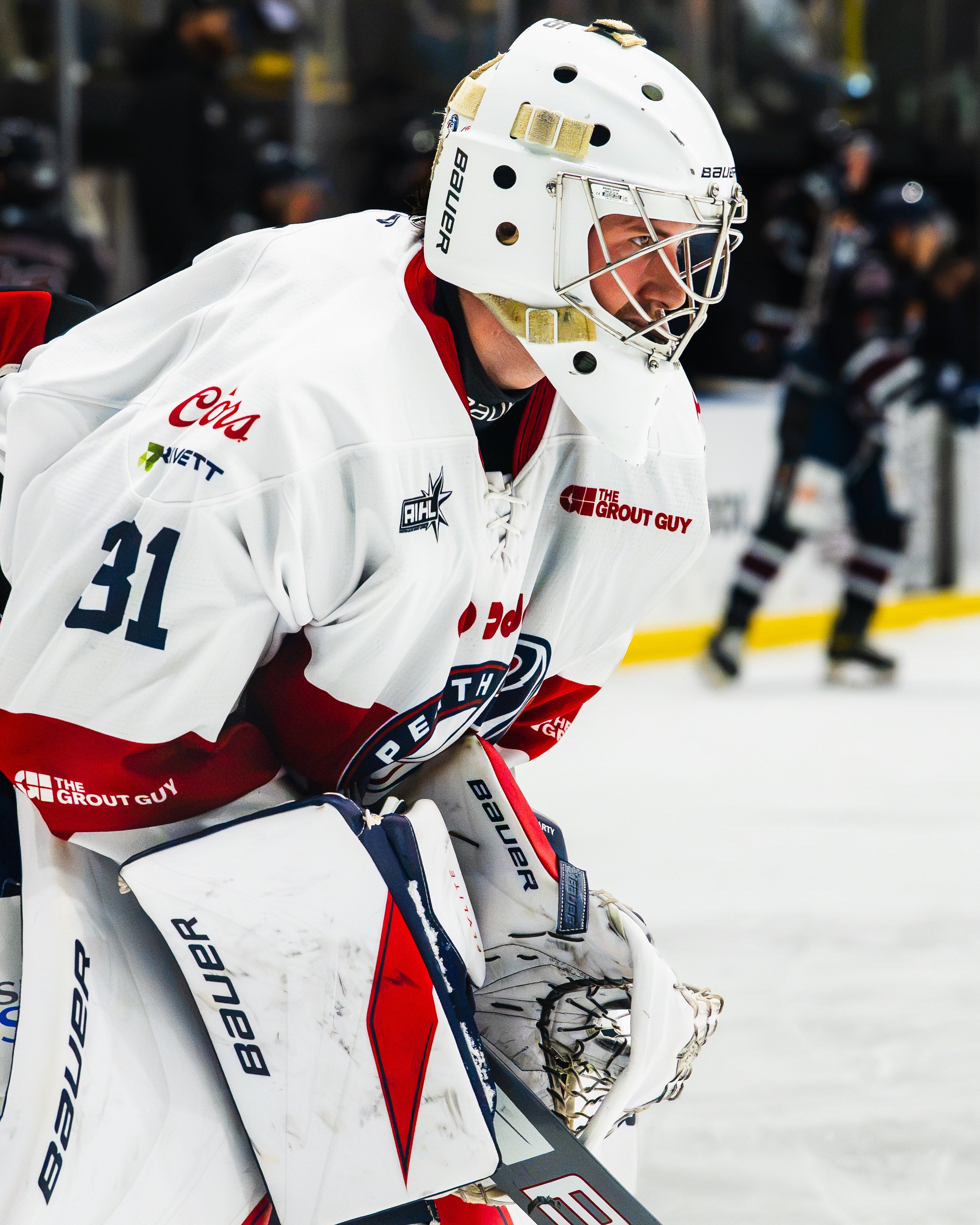 An ice hockey goalie in a white jersey and gear, wearing a helmet, crouches in the crease during a game, with spectators blurred in the background.