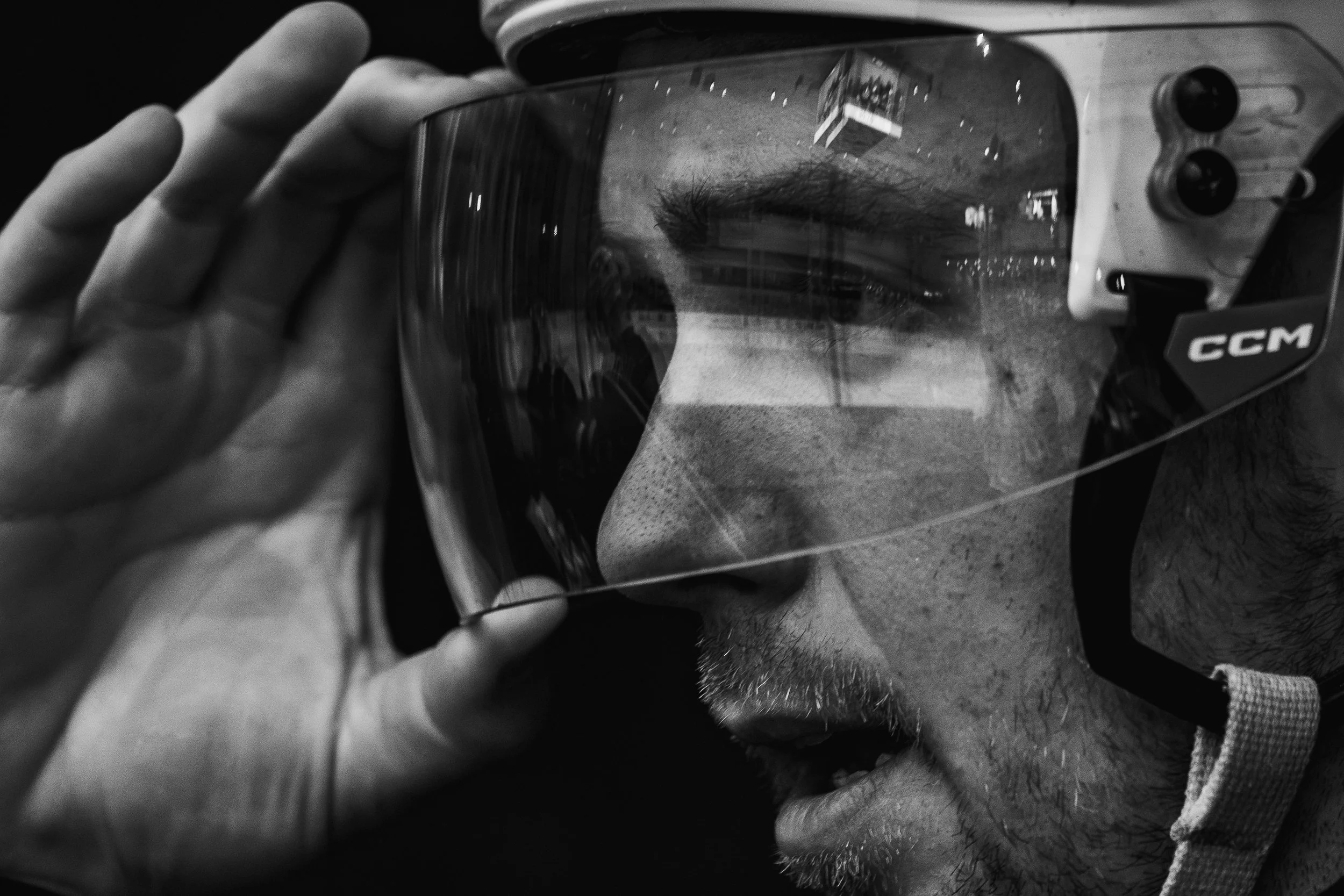 A person holding an ice hockey helmet with a reflective visor, and a reflection of a face with a beard in the visor. The person's hand is visible, and they are wearing a fingerless glove.