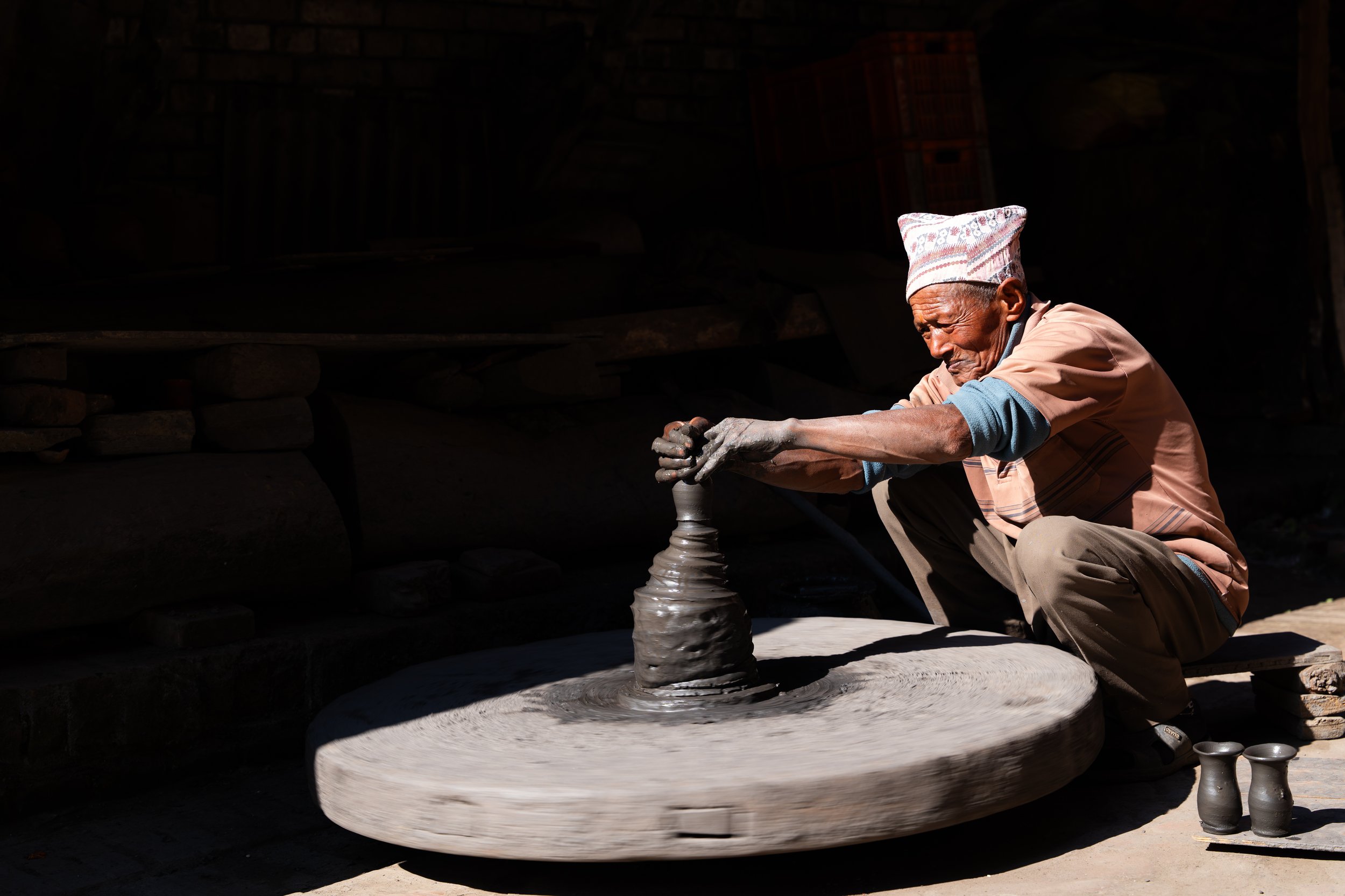 An elderly man wearing a traditional patterned hat and beige clothing kneels on the ground, shaping a large, circular piece of clay into a pot using a pottery wheel.