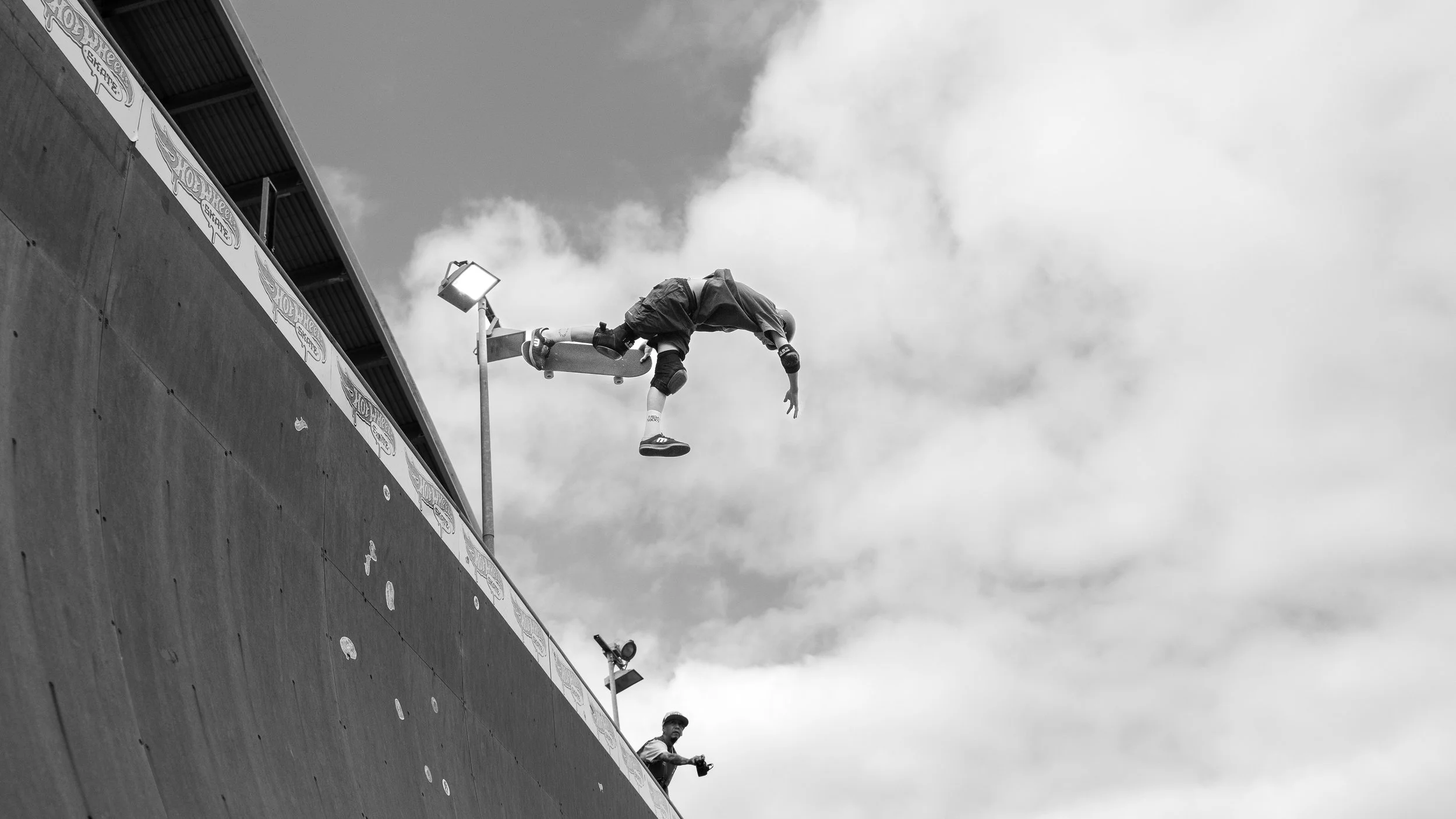 A skateboarder is mid-air performing a trick off a ramp at an outdoor skate park during the daytime, with a person holding a camera in the background and a cloudy sky overhead.