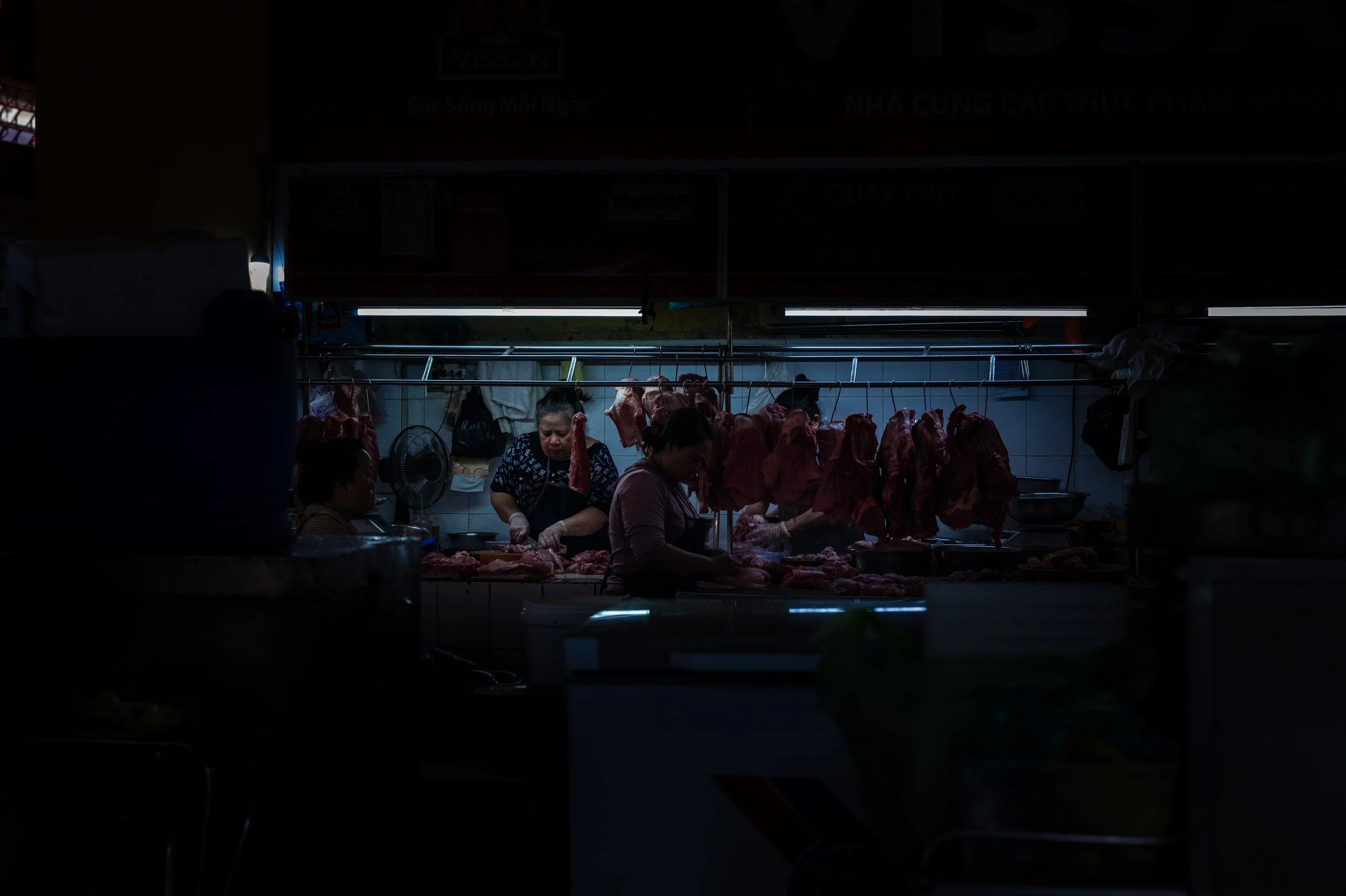 People working at a meat stall, hanging raw meat and preparing it, in a dimly lit indoor market.
