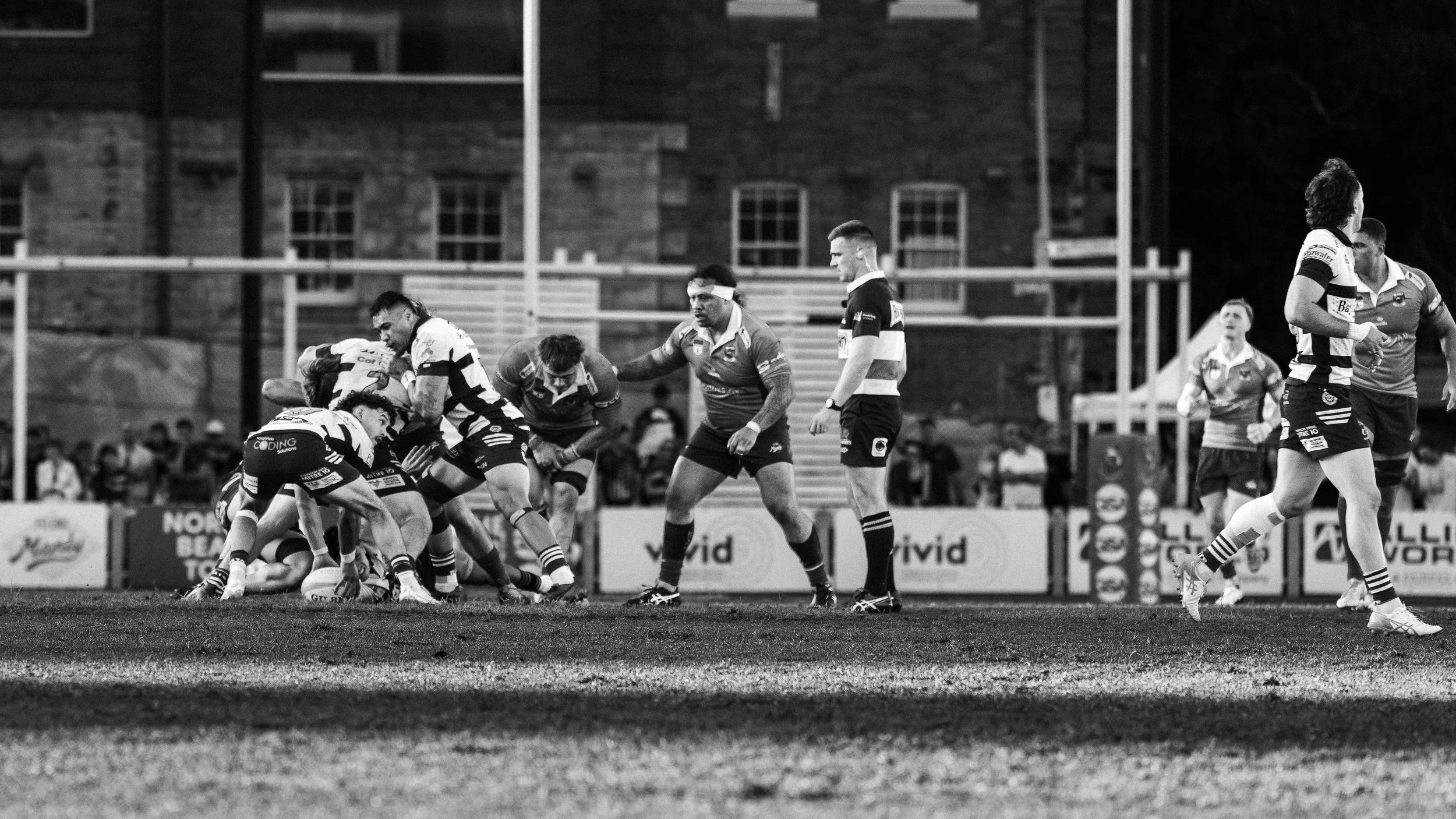 A black-and-white photo of a rugby match with players engaged in a scrum on the field, with spectators in the background.