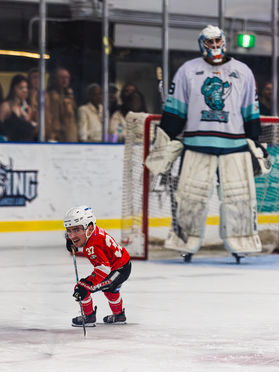 Young hockey player in red uniform skating on ice with a playful expression during a game, with a goalie and spectators watching in the background.