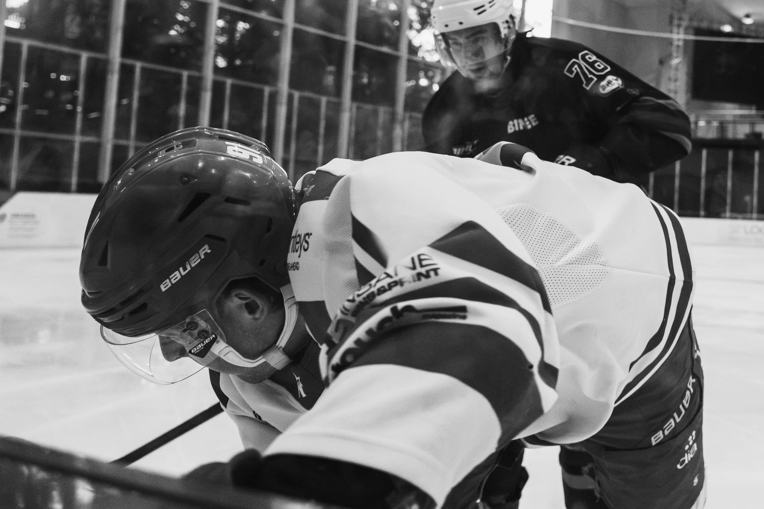 An ice hockey player in a helmet and uniform bent over on the ice, possibly during a game or practice, with another person in the background.