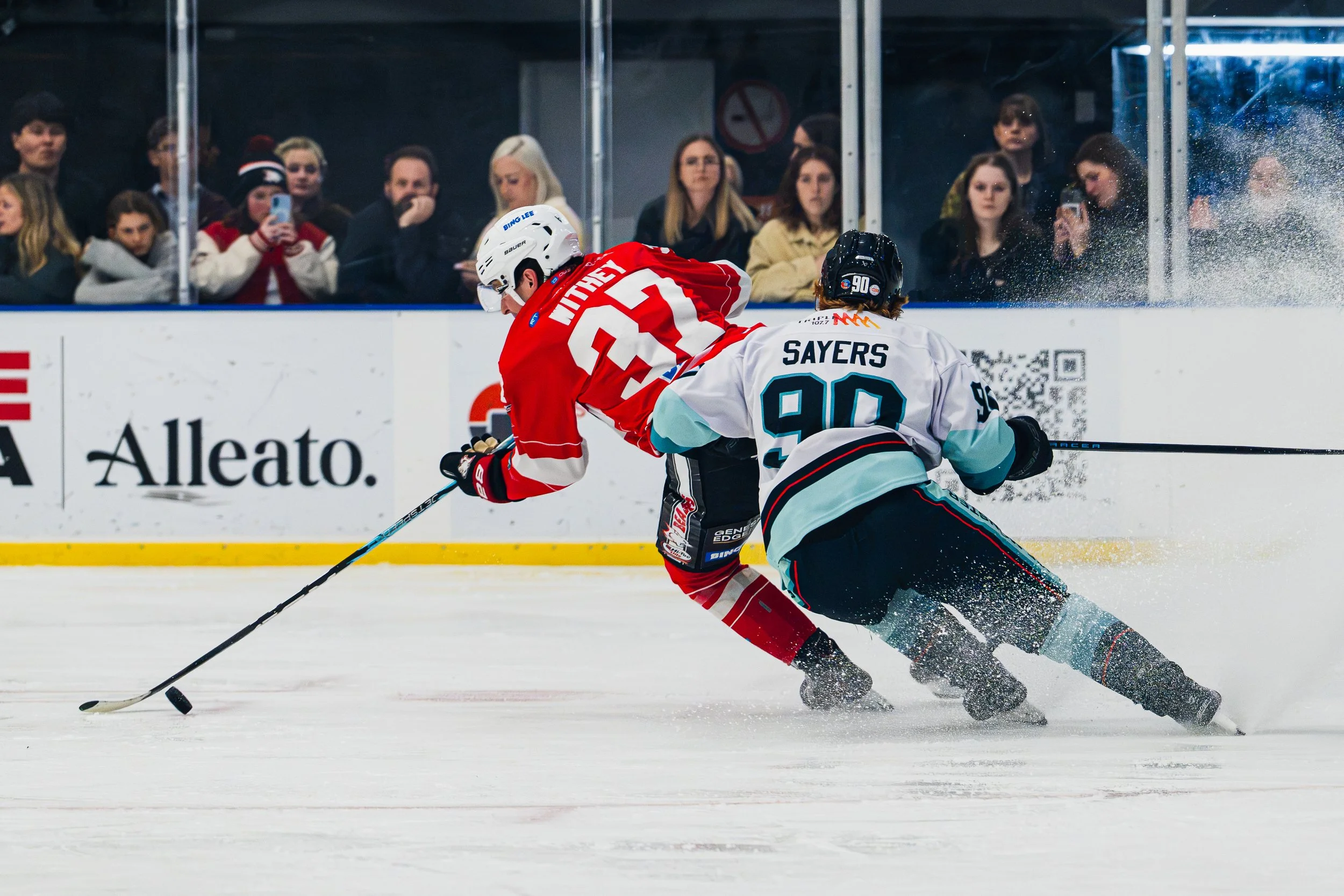 An ice hockey game with players in red and white jerseys skating on the ice. The player in the red jersey is hitting the puck, while the player in the white jersey, number 90, is trying to block. Spectators in the background watch the game, some taki