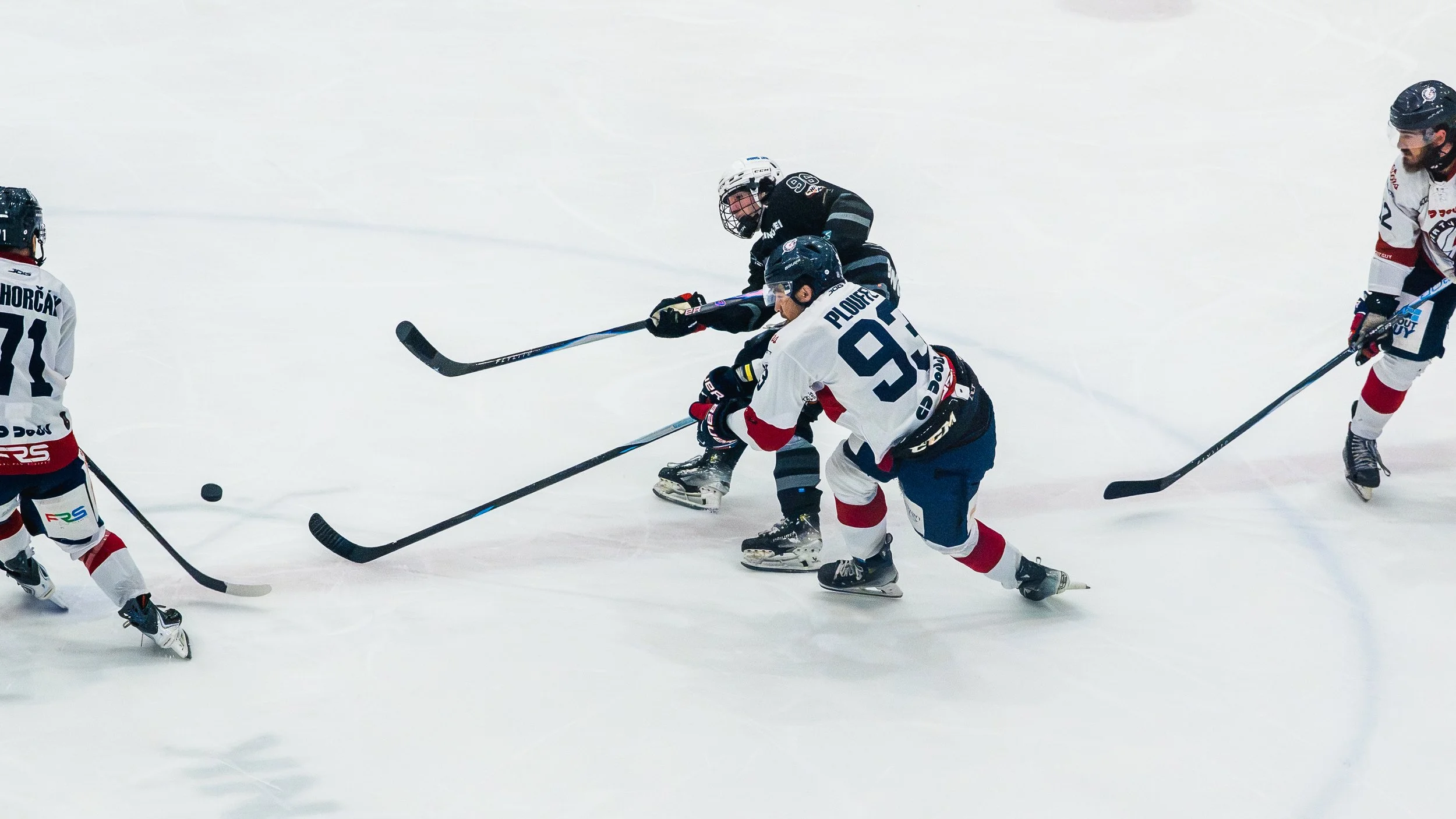 Ice hockey game with four players on the ice, two in white jerseys and two in black jerseys, actively pursuing the puck.