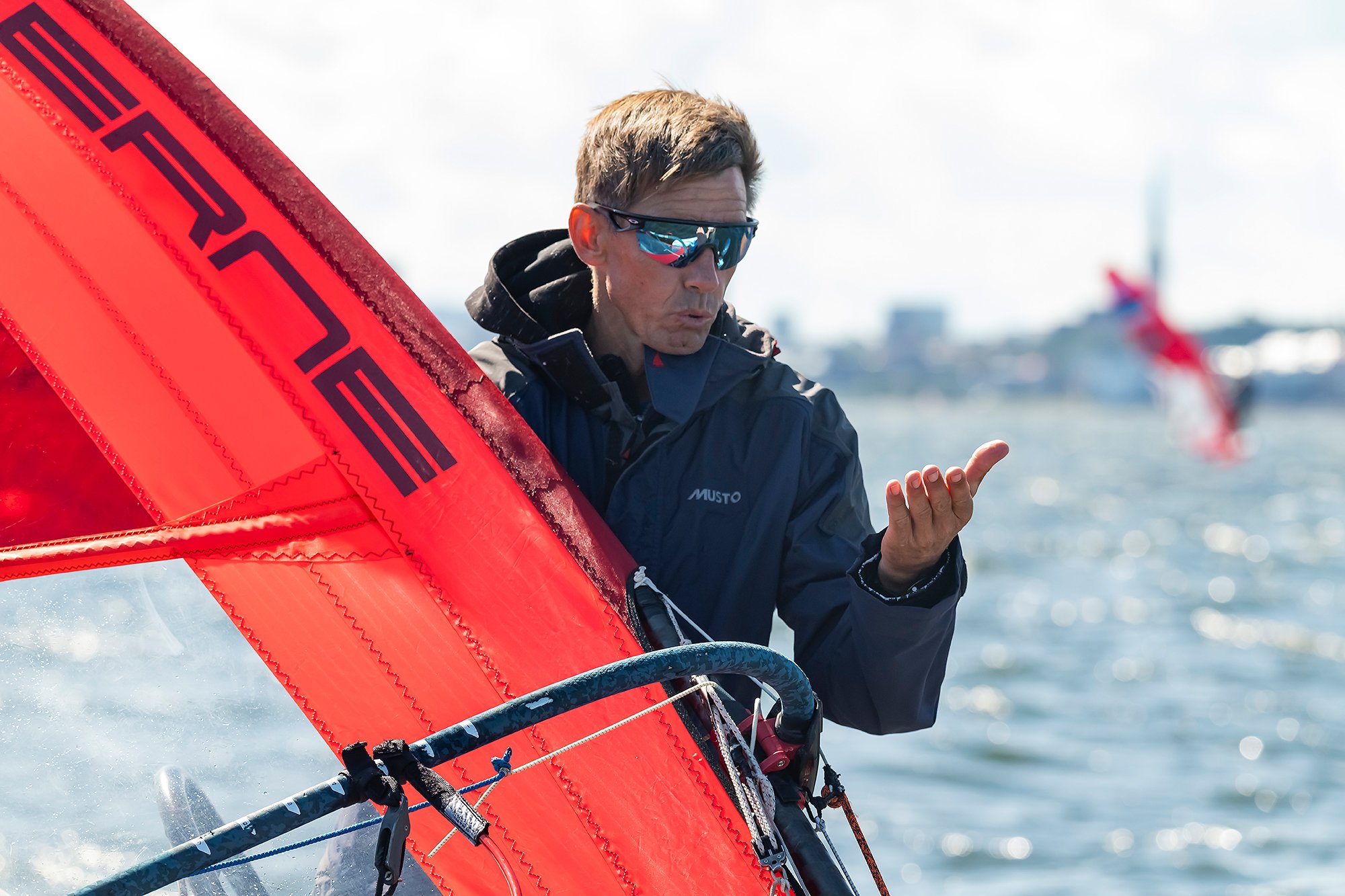 Man wearing sunglasses and a black jacket operating a windsurfing sail on the water with an airplane in the background.