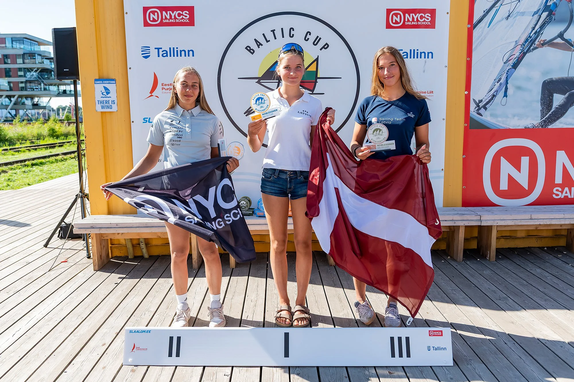 Three young women standing on a winners' podium after a sailing competition, holding their flags and medals, with a backdrop displaying the Baltic Cup logo and sponsor logos, at an outdoor location on a sunny day.
