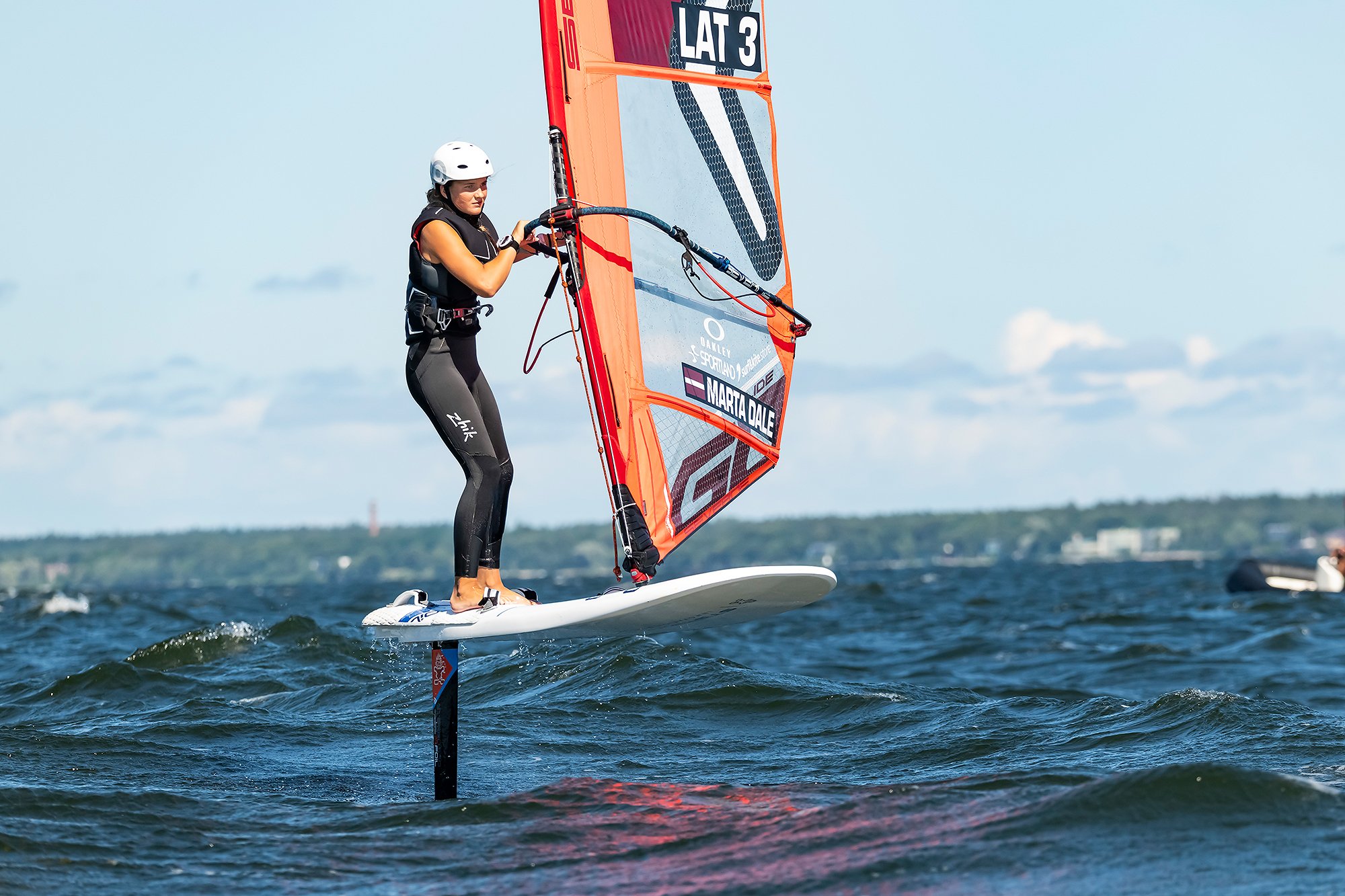 A woman windsurfing on a body of water, wearing a white helmet and black wetsuit, holding onto the sail of her windsurfing board.