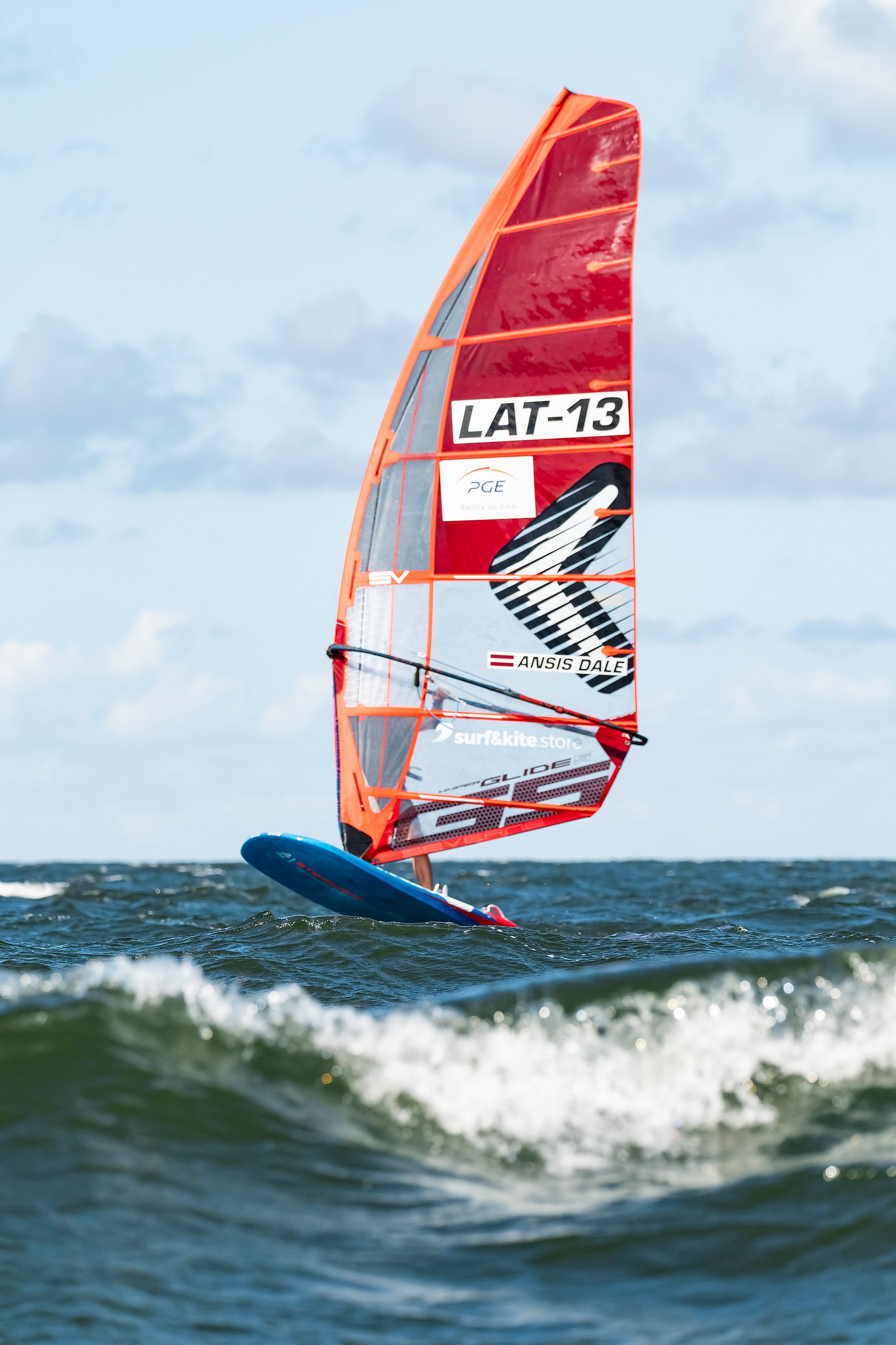 A person windsurfing on the water with a large red sail. The sail has branding and the sky is partly cloudy.