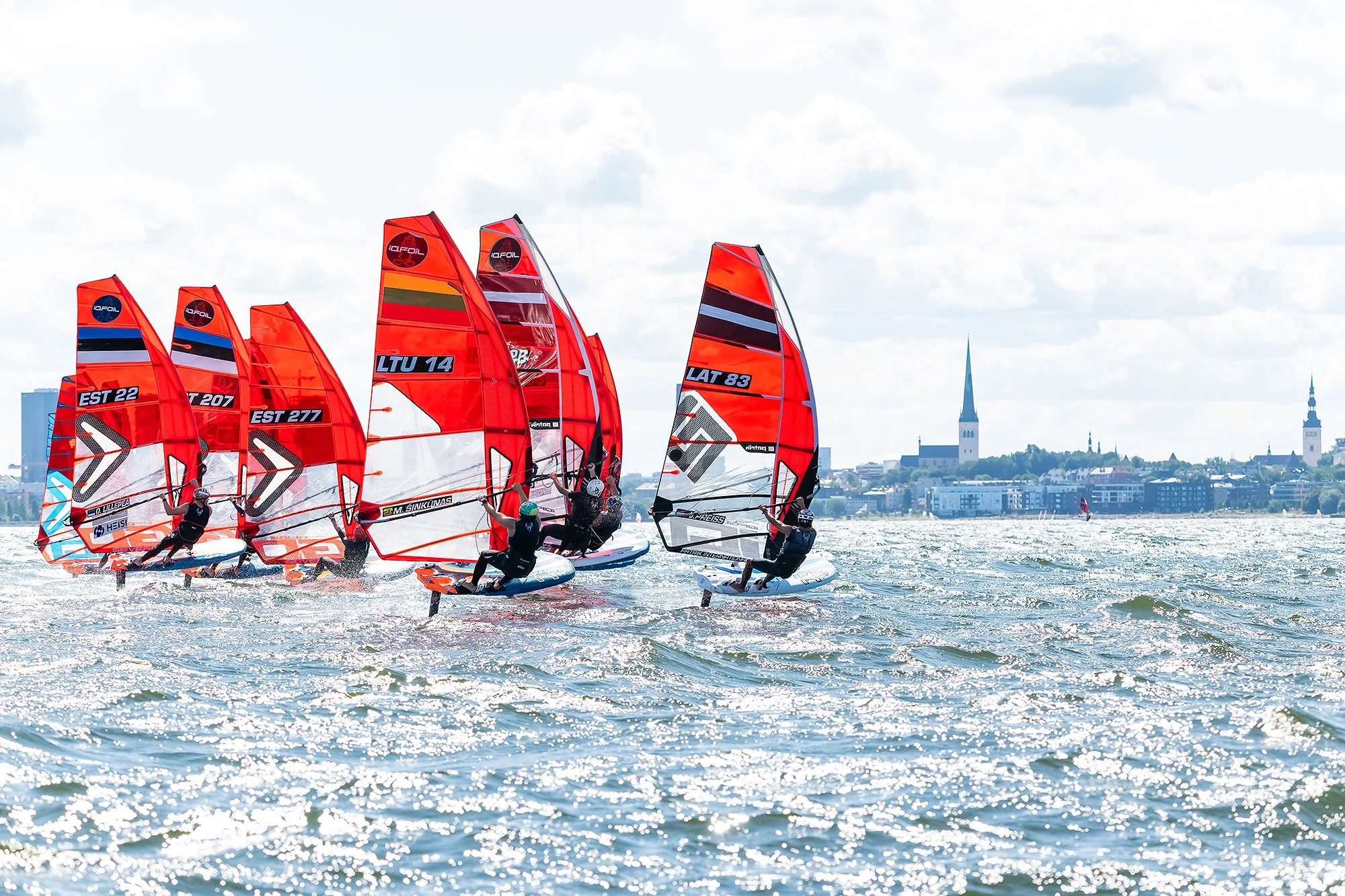 A group of windsurfers racing on the water with cityscape and church spires in the background.