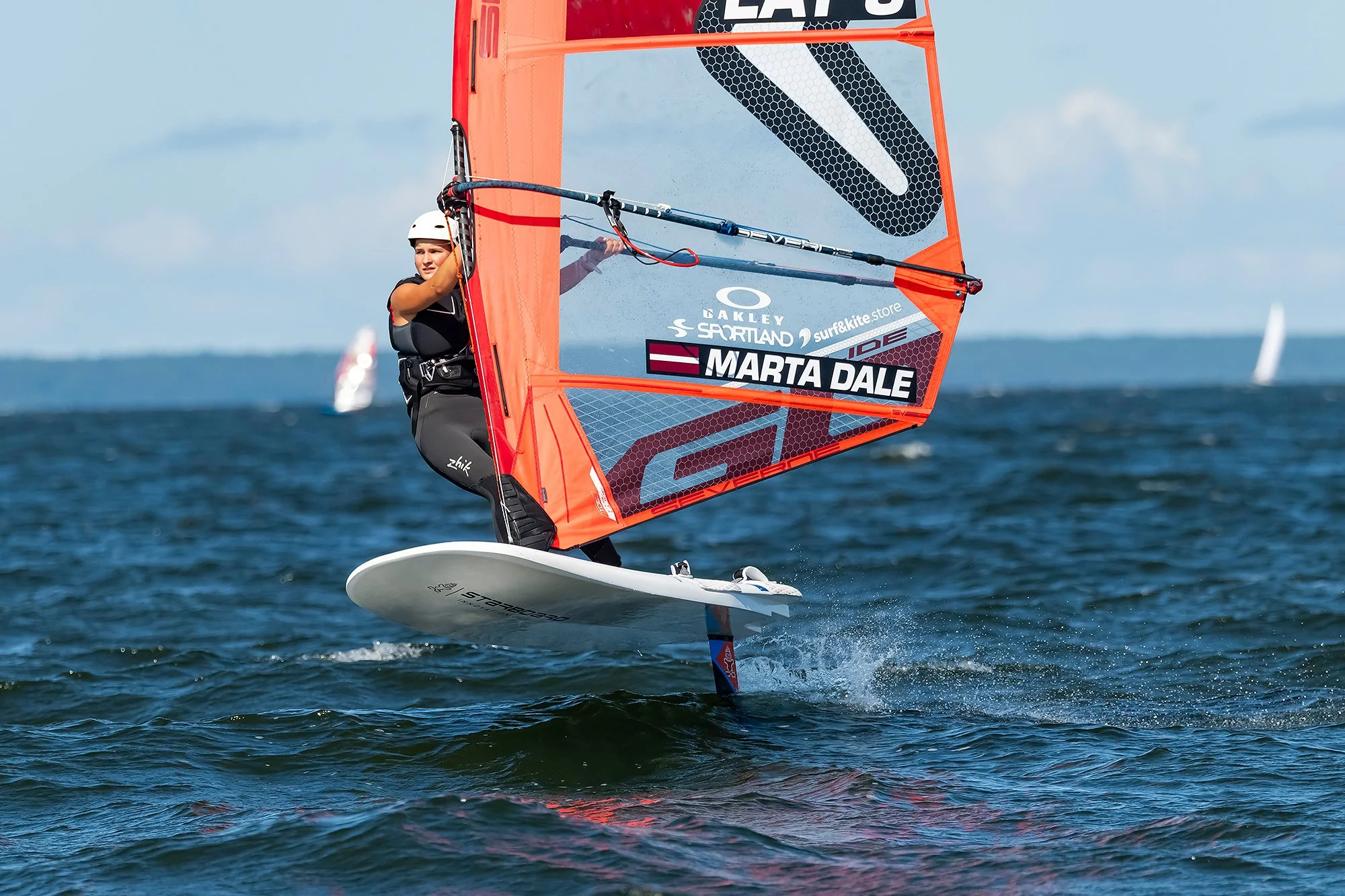 A woman windsurfing on the water, wearing a helmet and a life vest, holding onto the sail's boom as she rides a white board with a large orange sail that has the name 'Marta Dale' printed on it, with other sailboats visible in the background.
