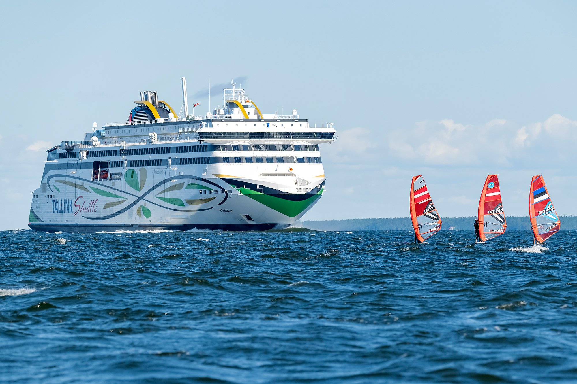 A large white cruise ship with colorful decals sailing on the water, with three red windsurfing sails and windsurfers in the foreground.