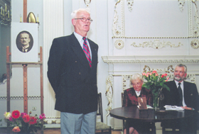Older man standing and speaking at a formal event in a decorated room with framed picture and floral arrangements.