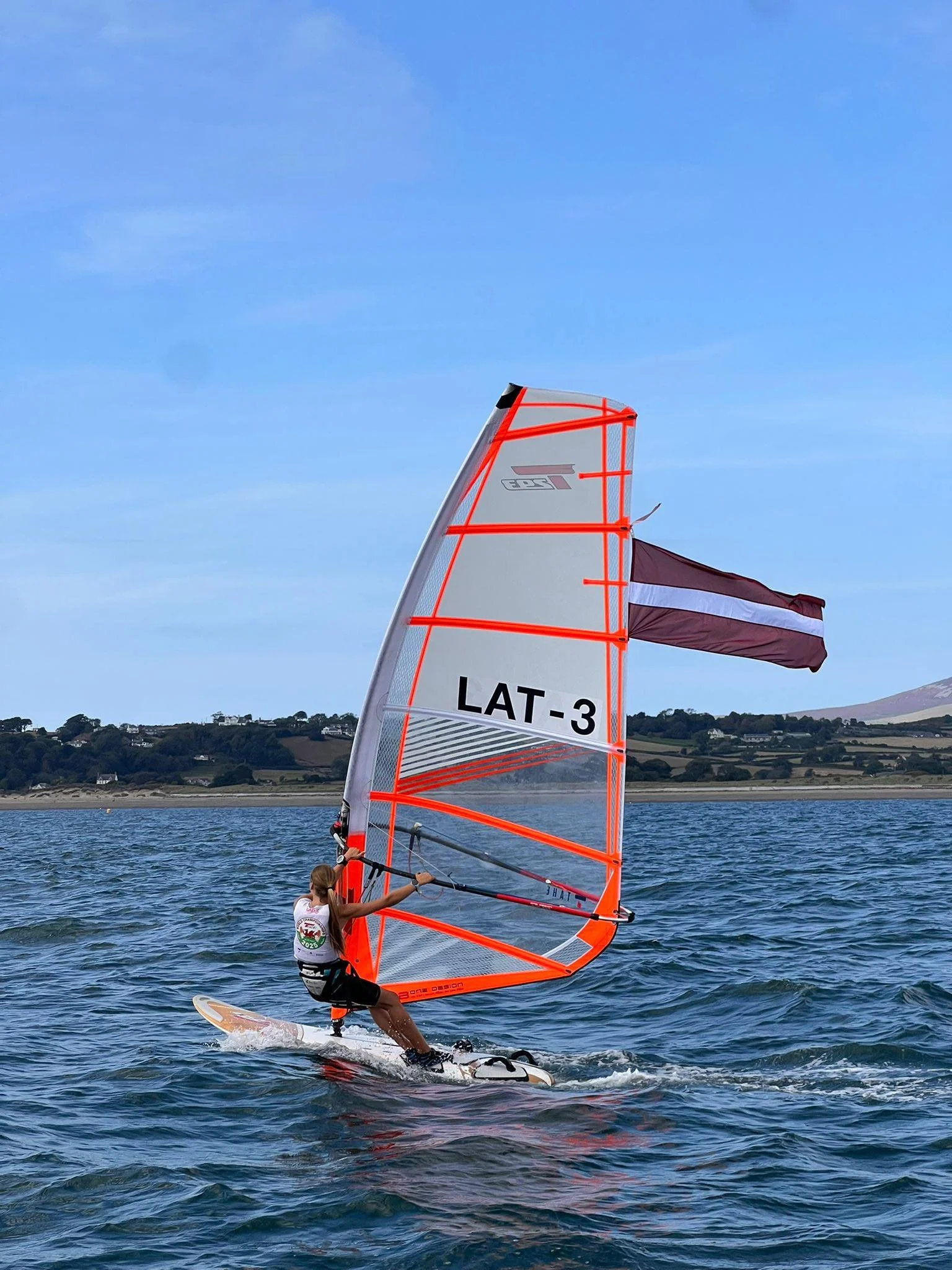 A person wearing a white shirt and black shorts sailing a small sailboat on a lake with hills in the background. The sailboat has a transparent sail with bright orange trim and the words "LAT-3" printed on it. The sky is clear with a few wispy clouds.