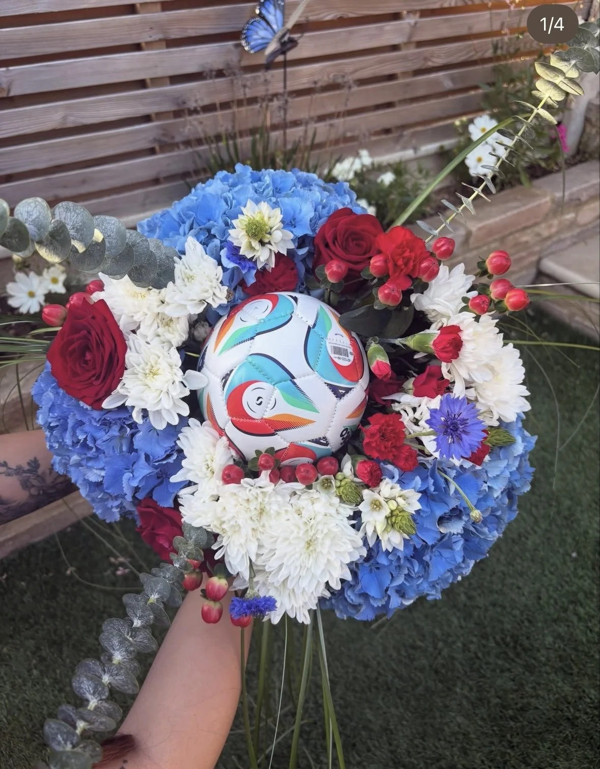 A bouquet of blue, red, white, and purple flowers with eucalyptus leaves, featuring a soccer ball with colorful patterns, held outside near a wooden bench.