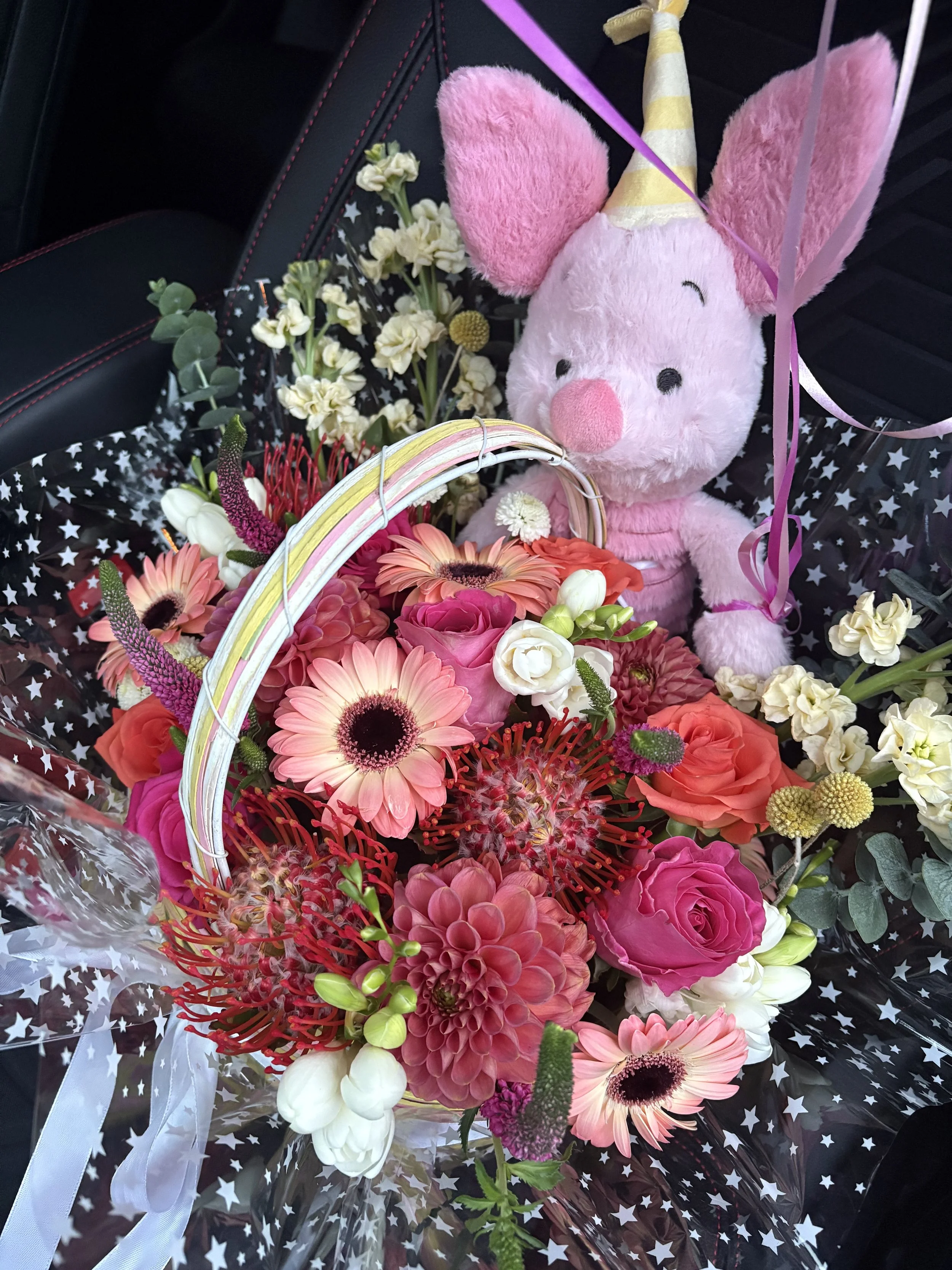 A pink plush stuffed animal with a unicorn horn and large pink ears surrounded by a colorful flower bouquet, wrapped in star-patterned cellophane.