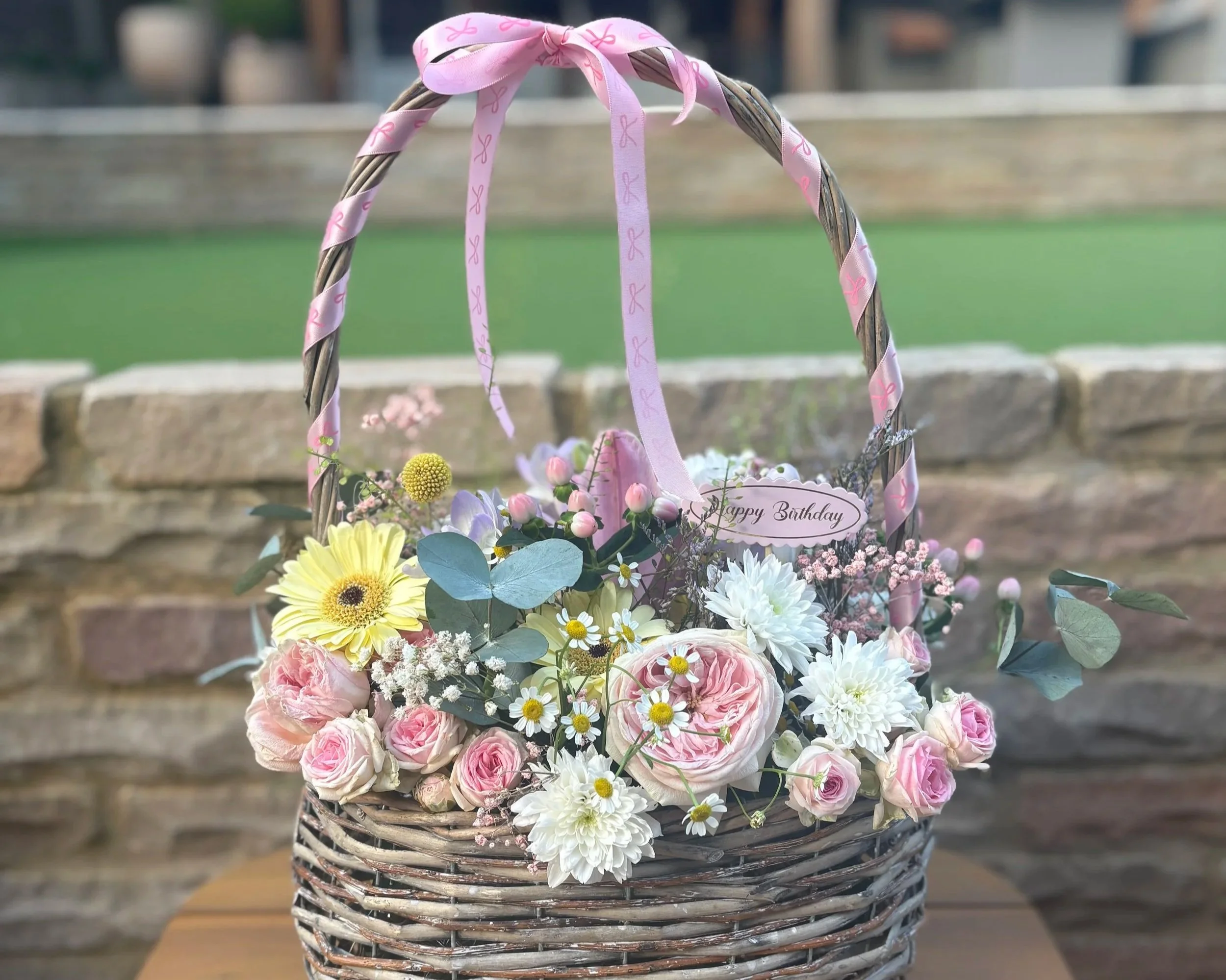 A wicker basket decorated with pink ribbons and filled with flowers, including daisies, roses, and other blooms, sitting outside on a wooden surface with a blurred background.