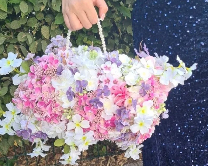 A person holds a handbag made of pink, white, purple, and pastel-colored flowers with a pearl-like handle, standing outside near green foliage.