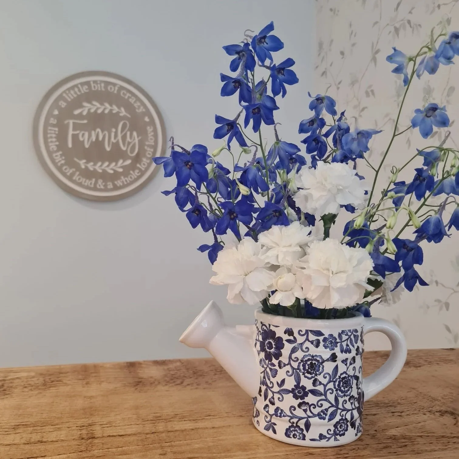 Blue and white floral arrangement in a decorative mug. Mug designed with blue floral pattern and serves as a vase. Background includes a wall sign with a quote and a wooden surface beneath the arrangement.