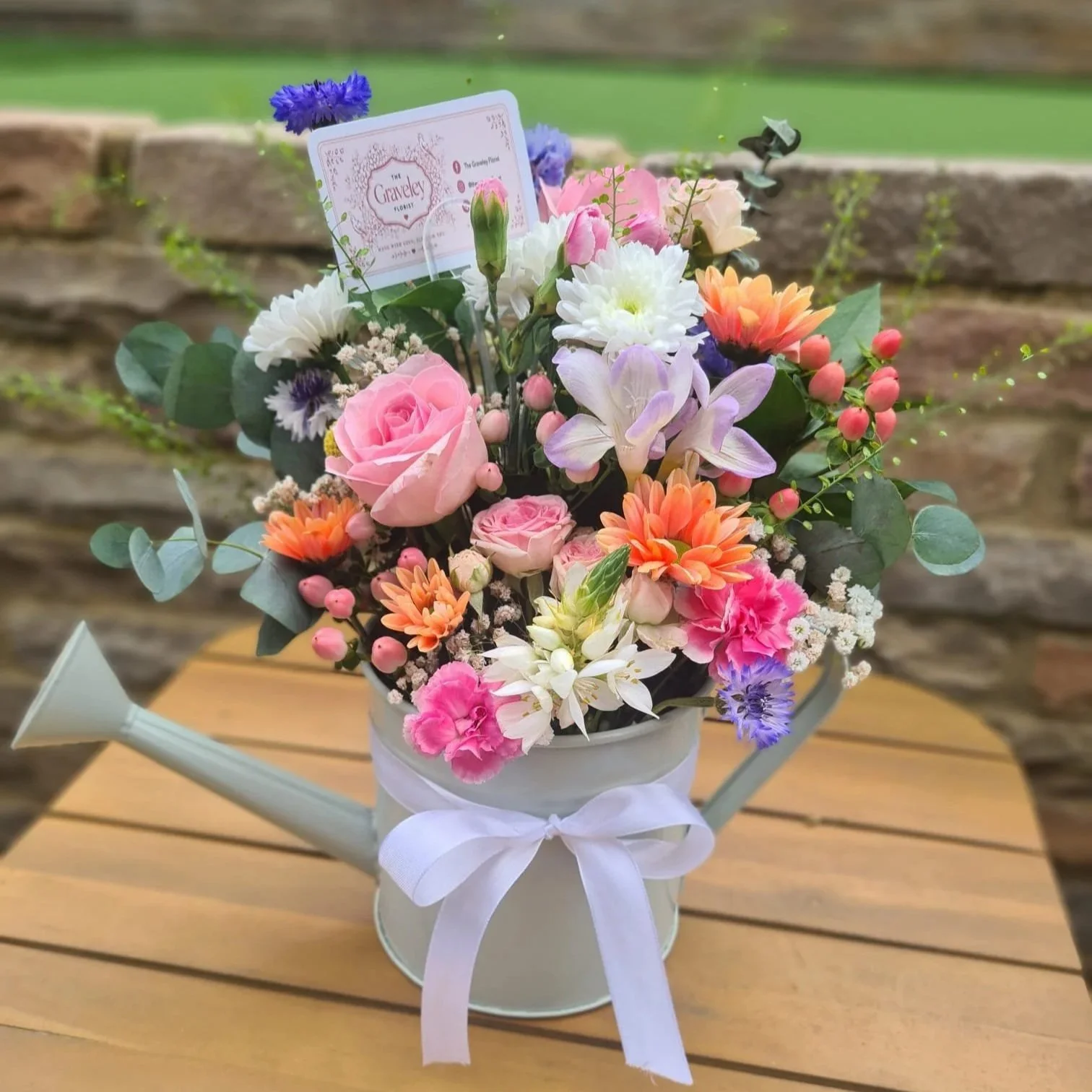 A white watering can with a ribbon tied around its neck, used as a vase for a colorful bouquet of flowers. The bouquet contains pink roses, white daisies, purple and orange flowers, pink berries, and eucalyptus leaves. The watering can is placed on a