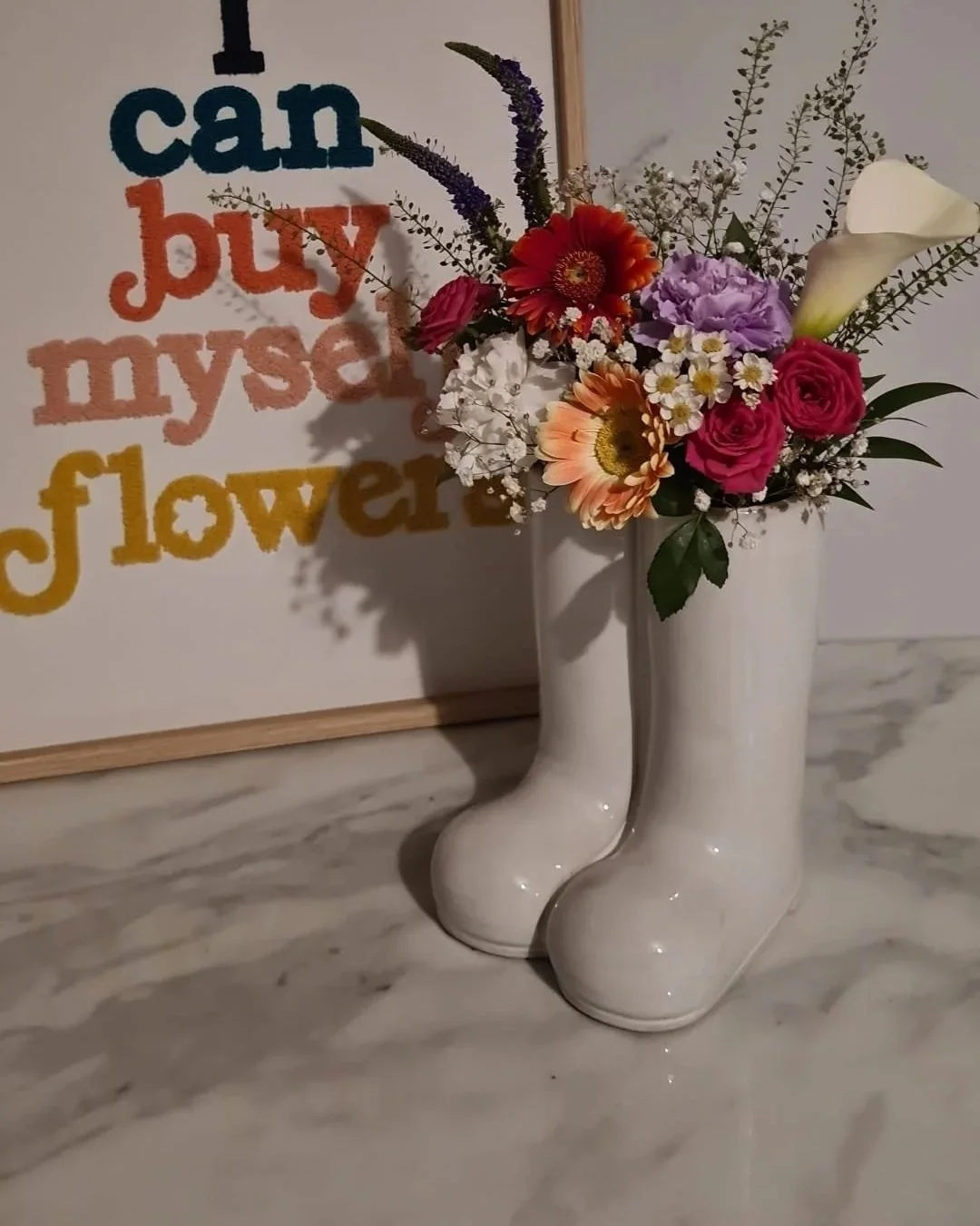A pair of white ceramic boots used as a vase, holding a colorful bouquet of flowers, next to a framed sign with the words 'I can buy my sweet flowers' in multicolor letters.